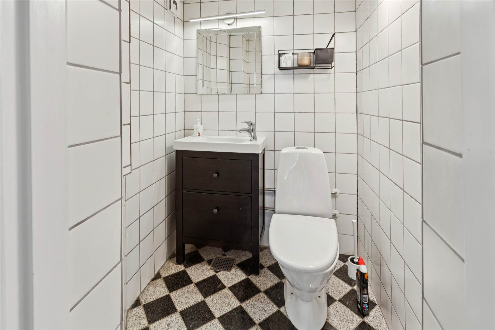 Et lille badeværelse med hvide vægge og et sort-hvidt gulv. En toilet, et hvidt toiletbord og en hængende hylde med lyseblå