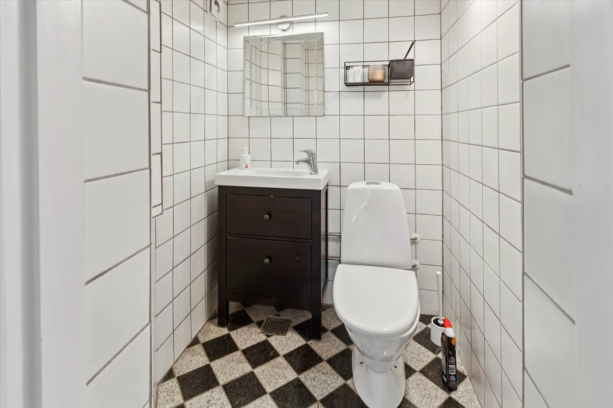 Et lille badeværelse med hvide vægge og et sort-hvidt gulv. En toilet, et hvidt toiletbord og en hængende hylde med lyseblå