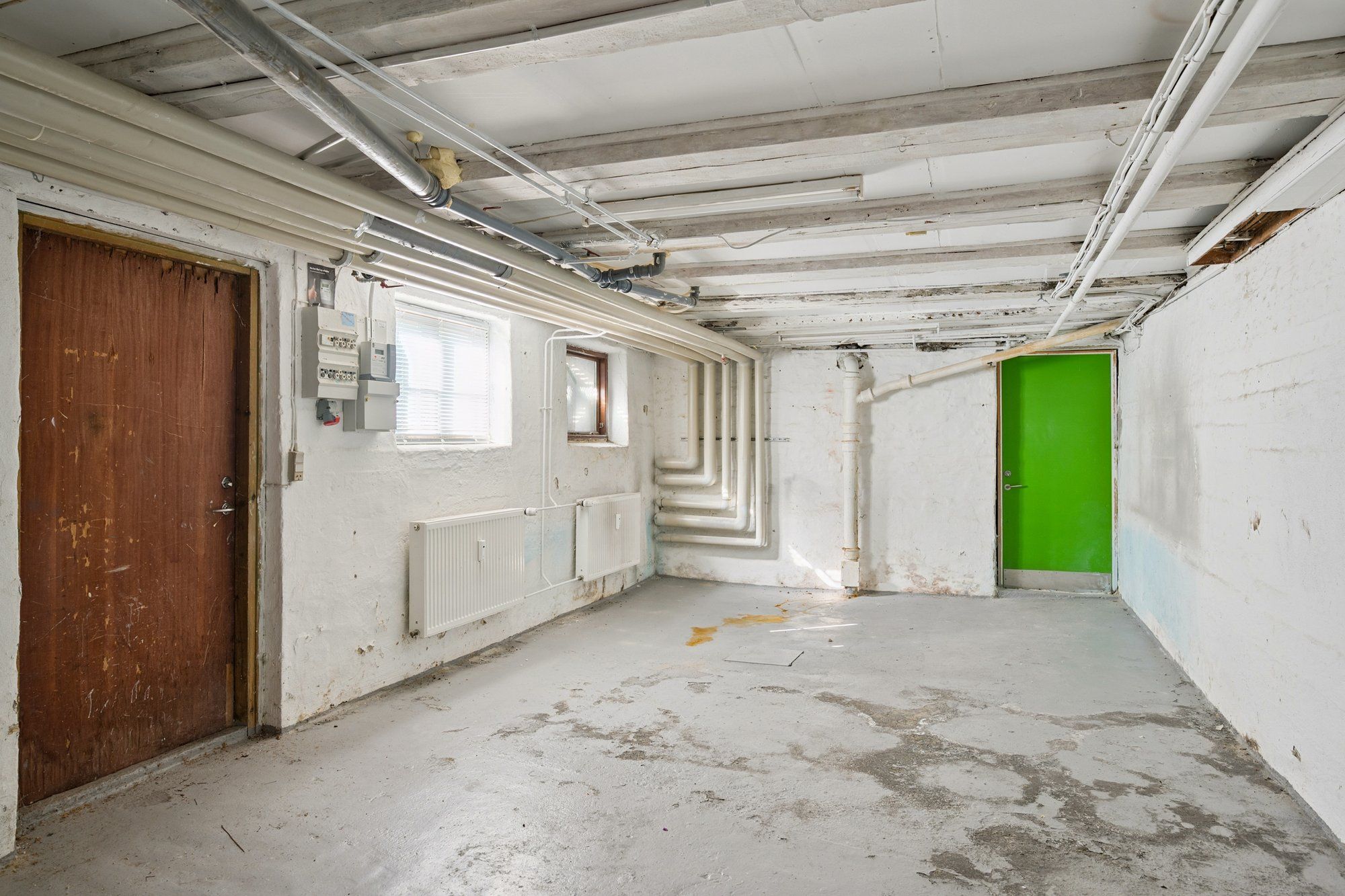 En kælder med to døre, den ene grøn og den anden brun, og hvide vægge med radiatorer og elektriske installationer
