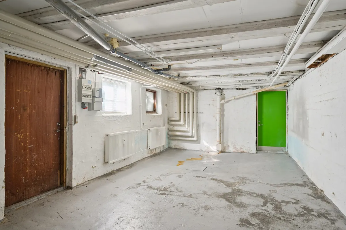 En kælder med to døre, den ene grøn og den anden brun, og hvide vægge med radiatorer og elektriske installationer