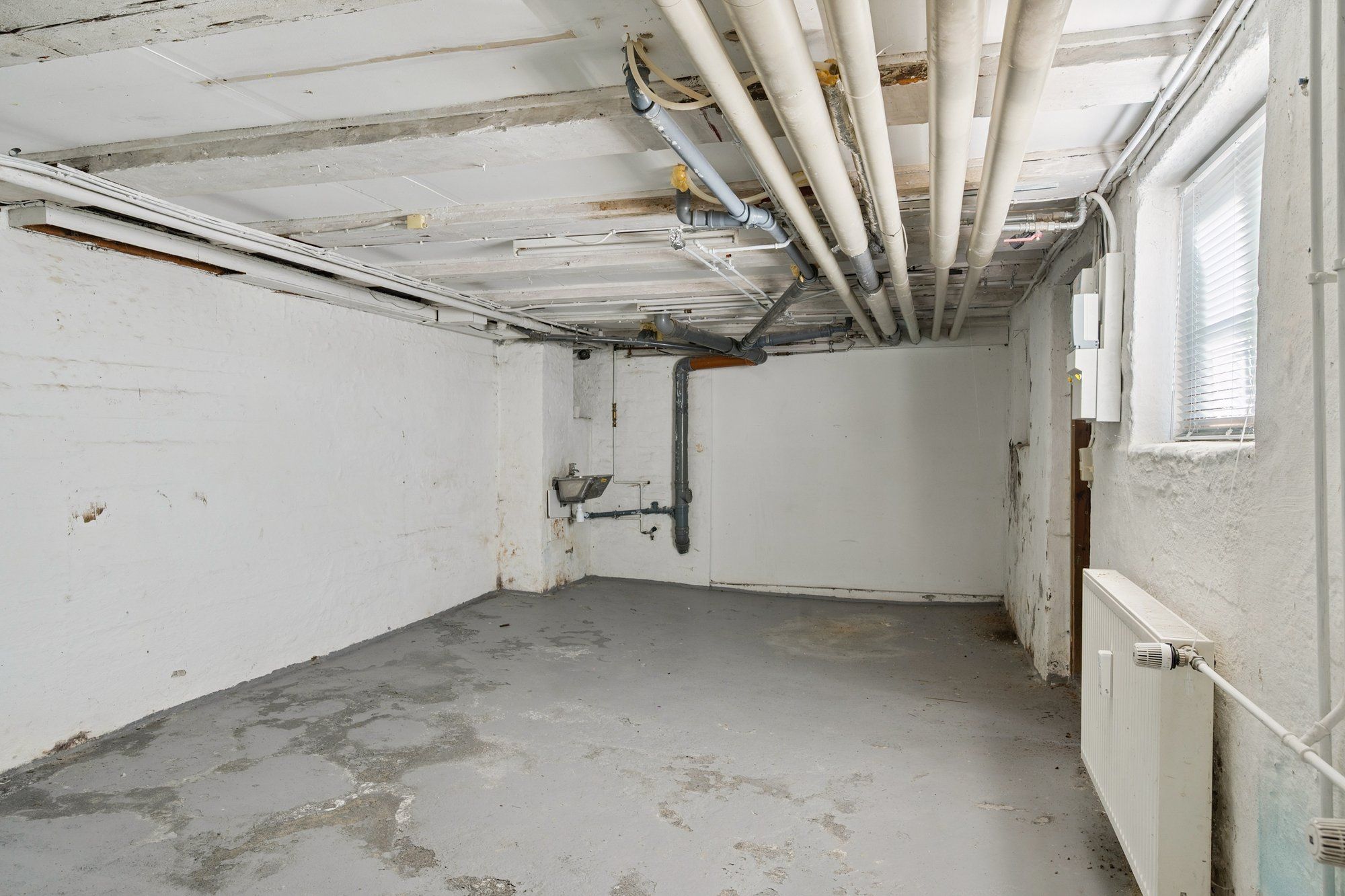 En kælder med hvide vægge og cementgulv, flere rør og kabel i loftet og en radiator i højre side