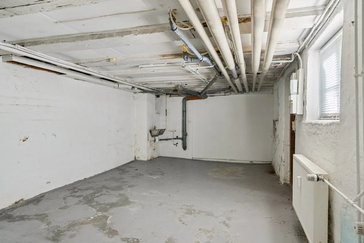 En kælder med hvide vægge og cementgulv, flere rør og kabel i loftet og en radiator i højre side