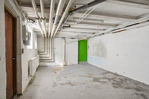 En kælder med hvide vægge og gulv. Her er en dør og et vindue. Væggen har et elektricitetspanel og rørene er hængende fra