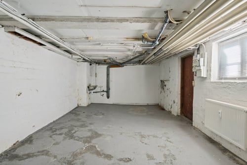 En tom kælder med hvide vægge og cementgulv, der ser ud til at være under renovering, med rør og elektriske ledninger i