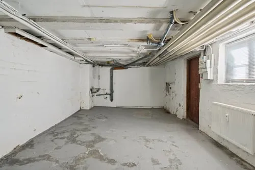 En tom kælder med hvide vægge og cementgulv, der ser ud til at være under renovering, med rør og elektriske ledninger i