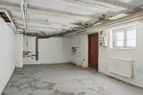 Lagerrum med hvidmalet væg, trægulv og piper samt radiator på den ene side og spildevandsrør på den anden.