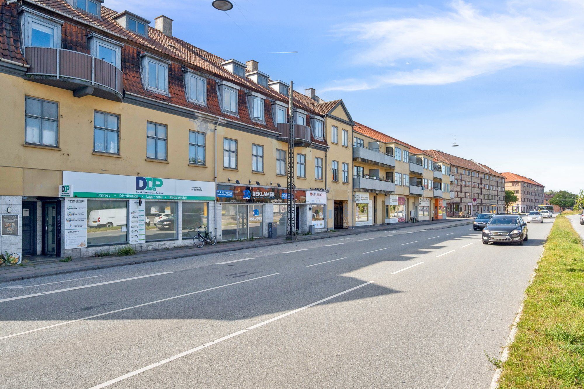 Et køretøj kører på en vej, der går gennem en by. Der er flere biler på vejen og nogle biler parkeret på siden af vejen. Der