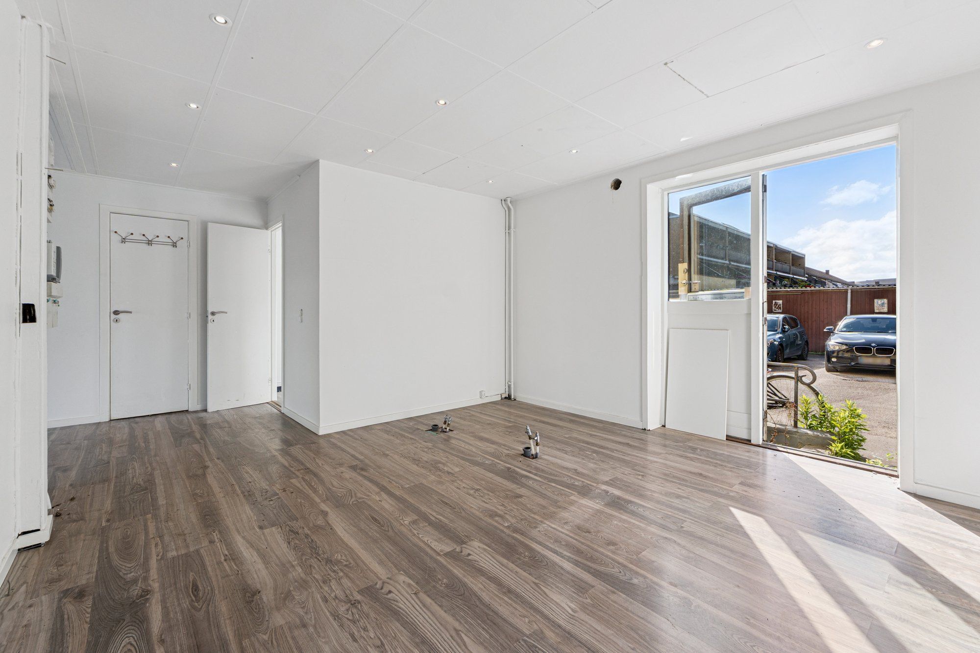 Minimalistisk indretning med lysegrå vægge og trægulve, åben dør og glasdør til gårdspladsen med parkerede biler og grønne