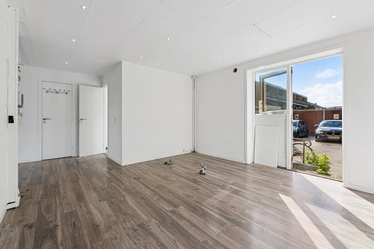 Minimalistisk indretning med lysegrå vægge og trægulve, åben dør og glasdør til gårdspladsen med parkerede biler og grønne