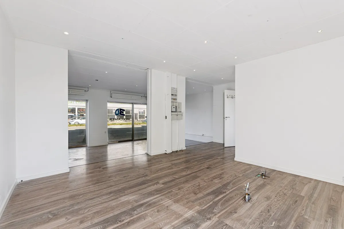 En stor, tom hvid stue med blanke gulvbelægning, store vinduer og åbne døre, der vender ud mod et parkeringsområde med biler