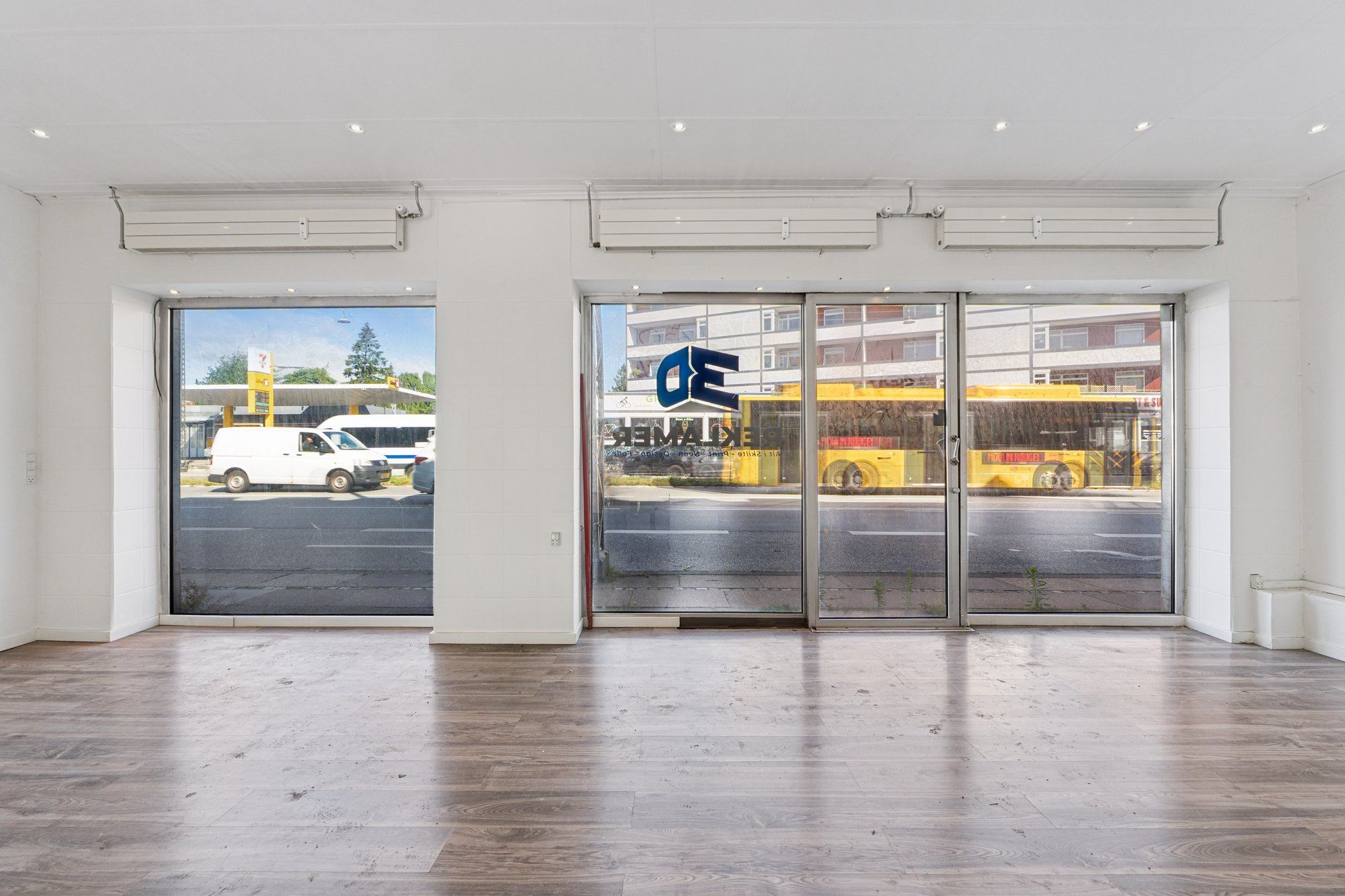 Store vinduer og døre til en butik med trægulv og hvide vægge med busser og en tankstation bagved