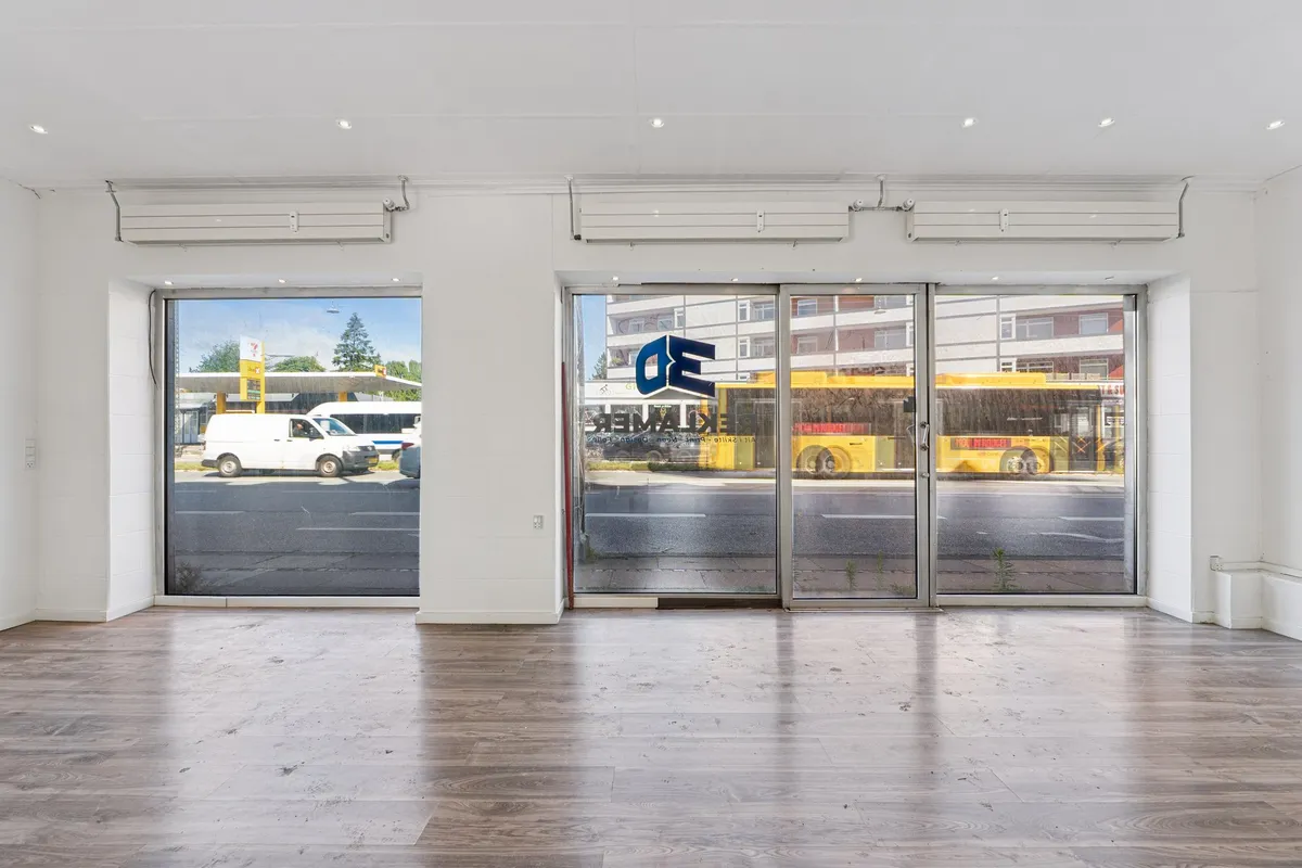 Store vinduer og døre til en butik med trægulv og hvide vægge med busser og en tankstation bagved