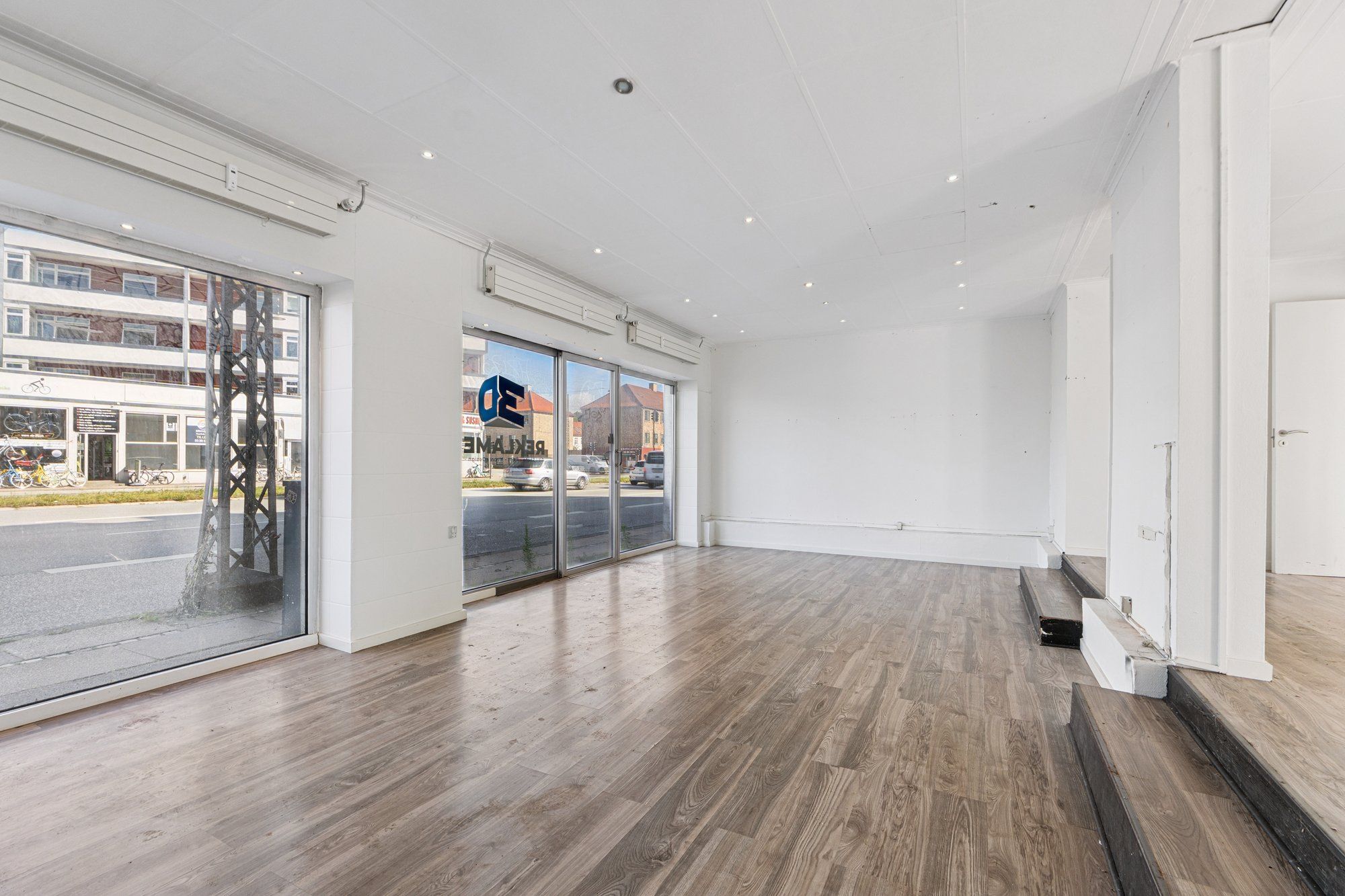 En tom stue med højloft, gulvet er i lysegråt træ og der er flere vinduer med glasdøre