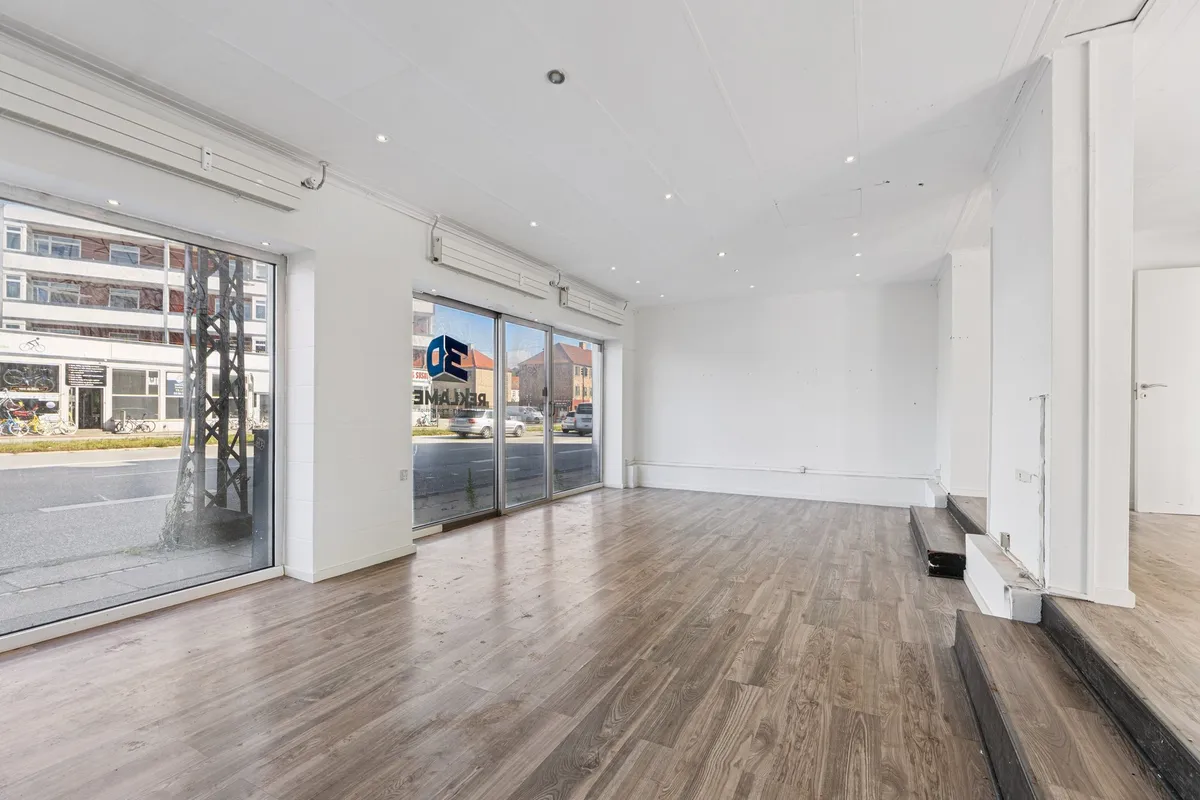 En tom stue med højloft, gulvet er i lysegråt træ og der er flere vinduer med glasdøre