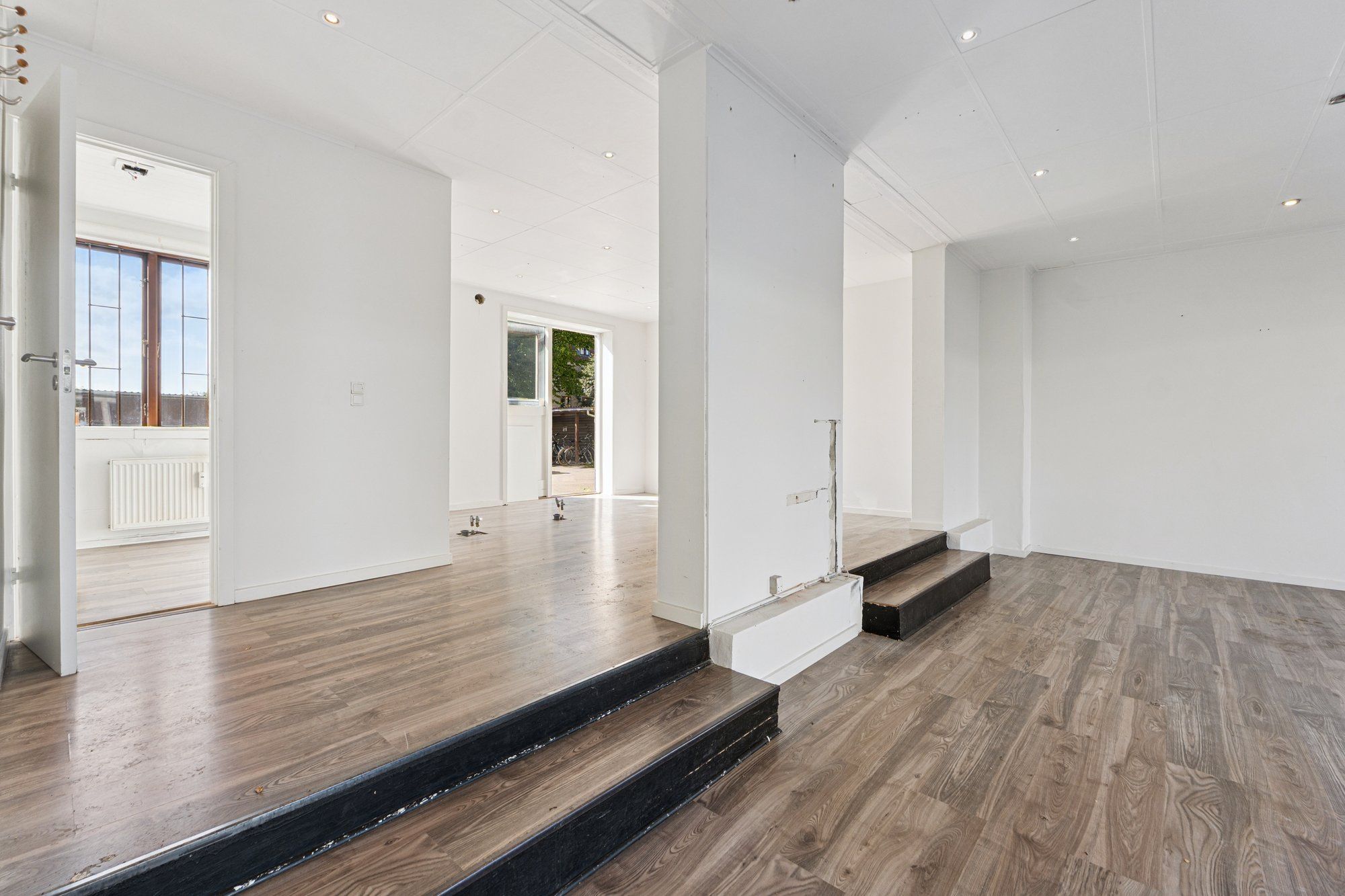 Interiør med hvide vægge, trægulve, åbne døre og høje lofter. Vinduesglas, radiator og stiger