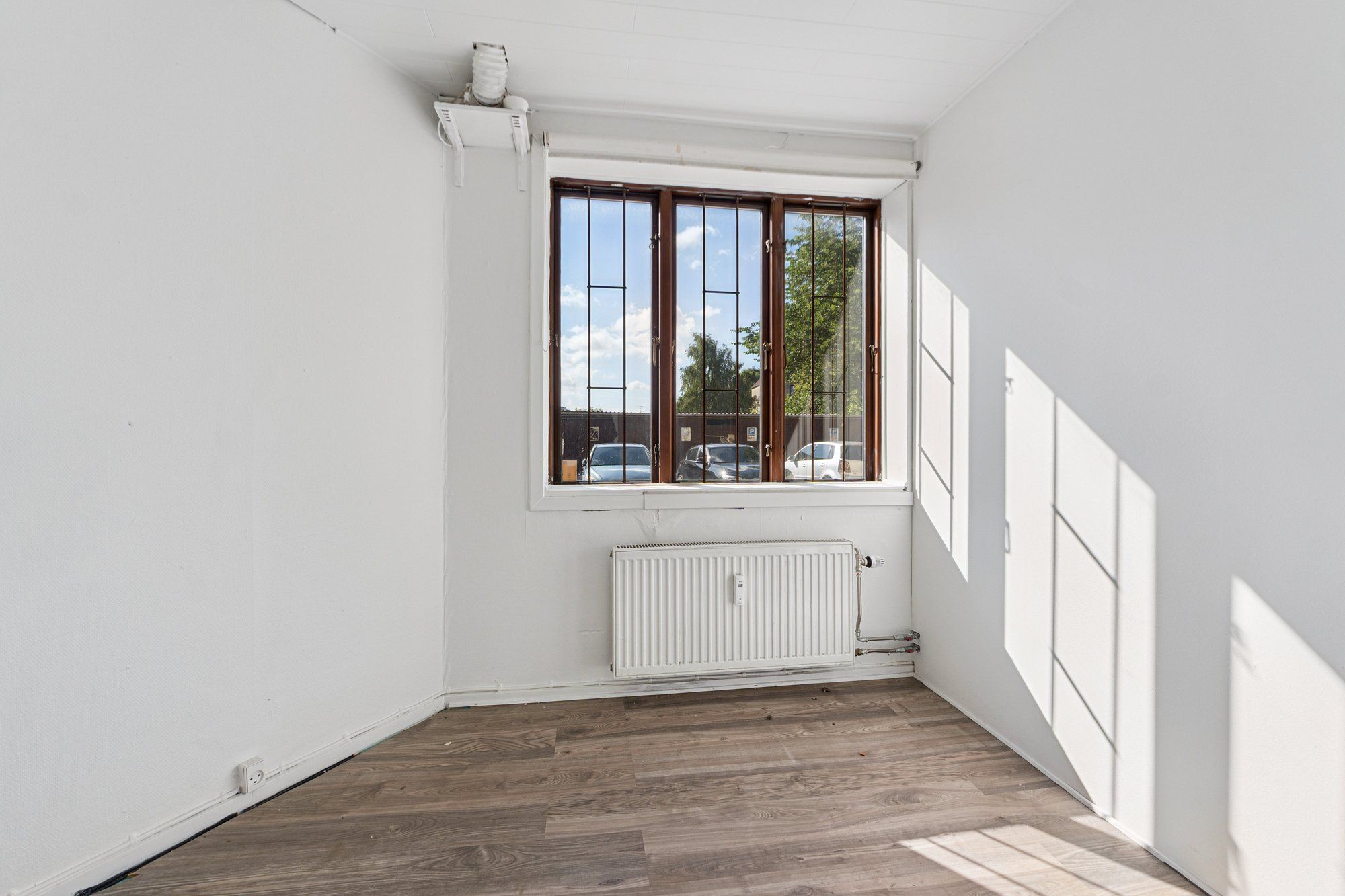 Et tomt værelse med hvide vægge og et gulv med træpaneler samt et vindue med metalruder og en radiator