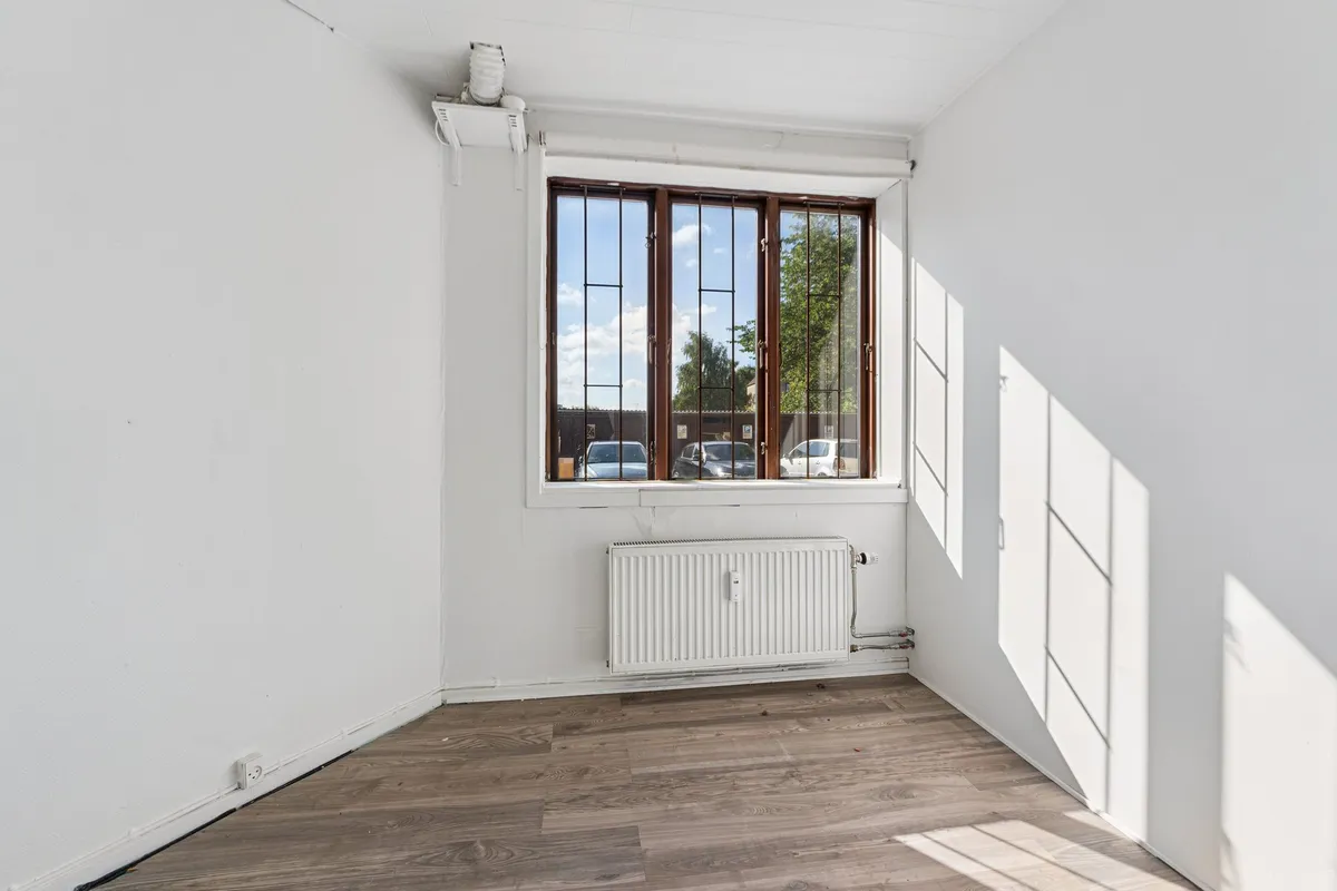 Et tomt værelse med hvide vægge og et gulv med træpaneler samt et vindue med metalruder og en radiator
