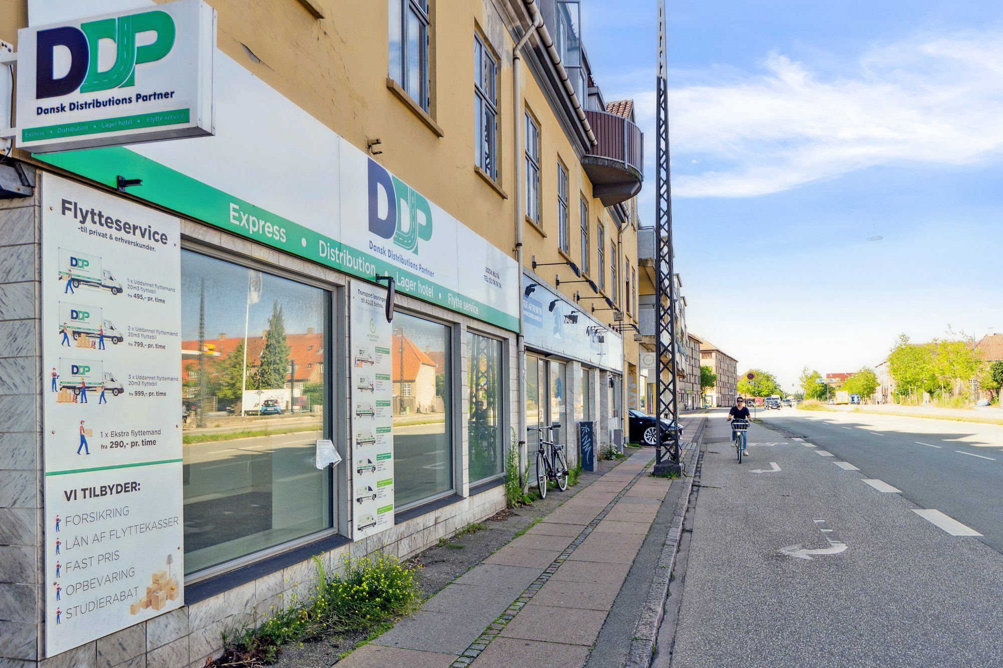 En kvinde kører på cykelstien med en cykel med kurv, mens hun passerer forretningen DDP med flytteservice, lagerhotel og