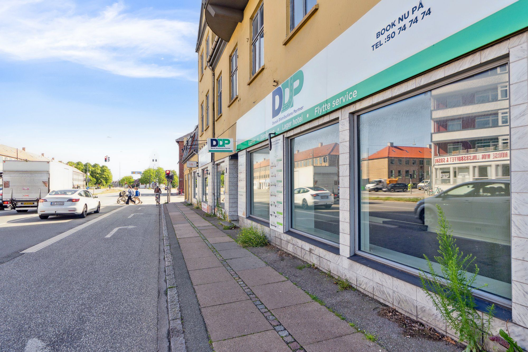 Butikslokaler med skrift og logoet DDP og flere biler kører forbi på en tosporet gade