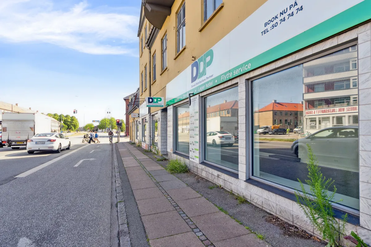 Butikslokaler med skrift og logoet DDP og flere biler kører forbi på en tosporet gade