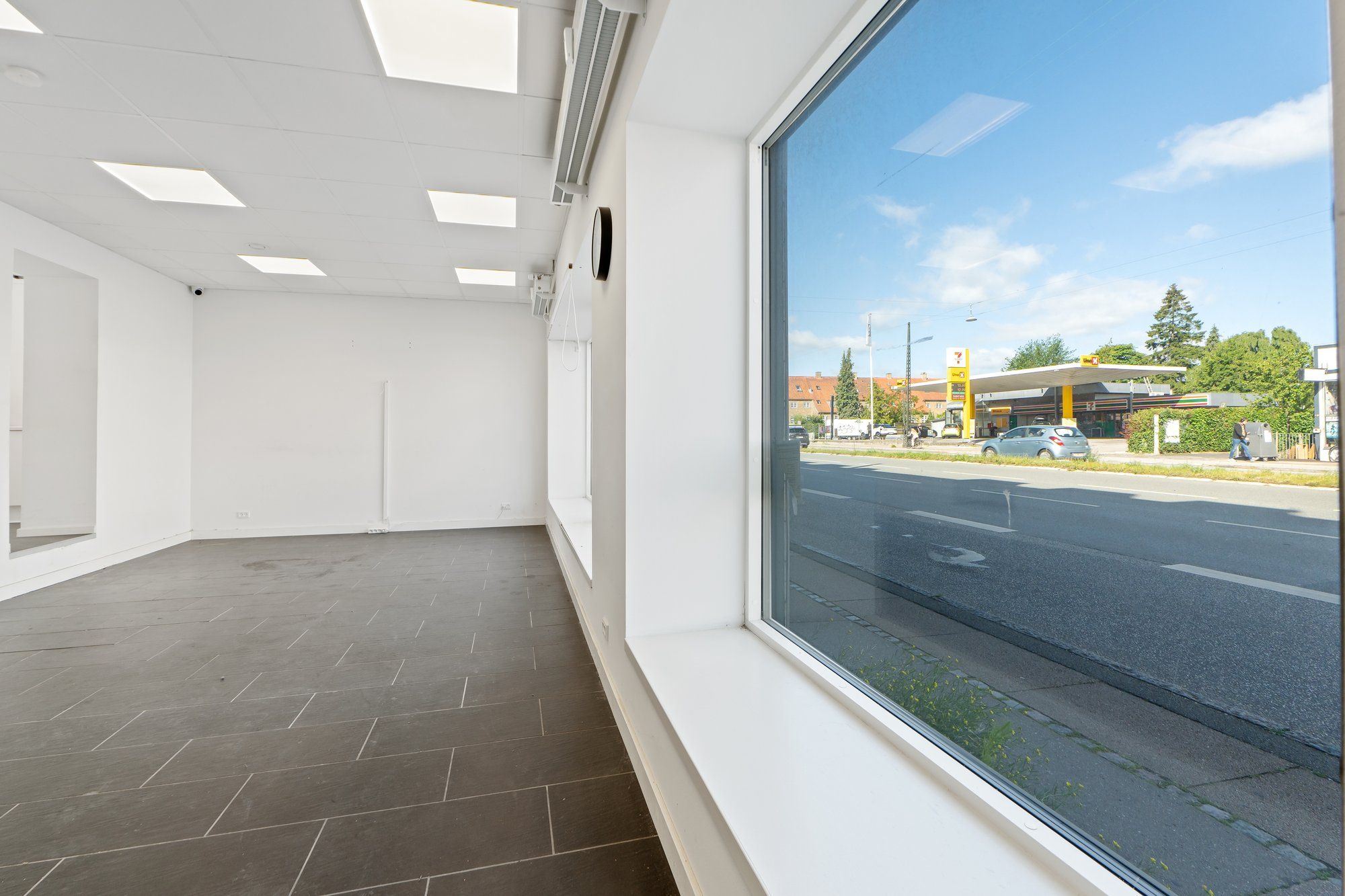 En stor glasrute vender ud mod en tosporet asfaltvej, hvor en bil kører langs med en benspærret fortov. Forruden af bilen