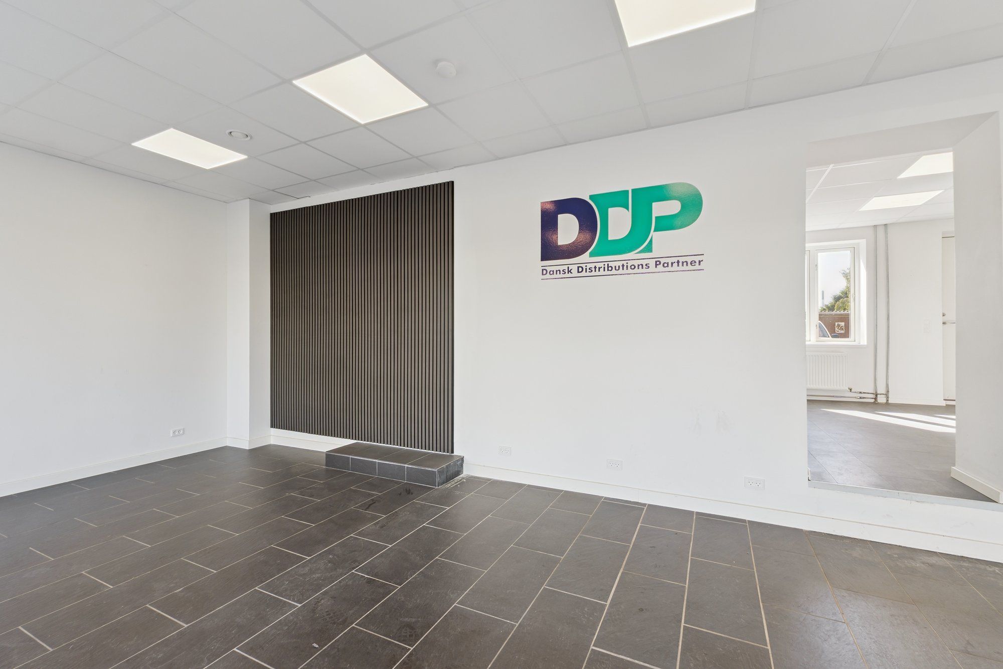 Indenfor et hvidt kontor med tavle med DDP tekst og logo og en sort skrå tavle. Gulvet er lysegrå og loftet er hvidt med