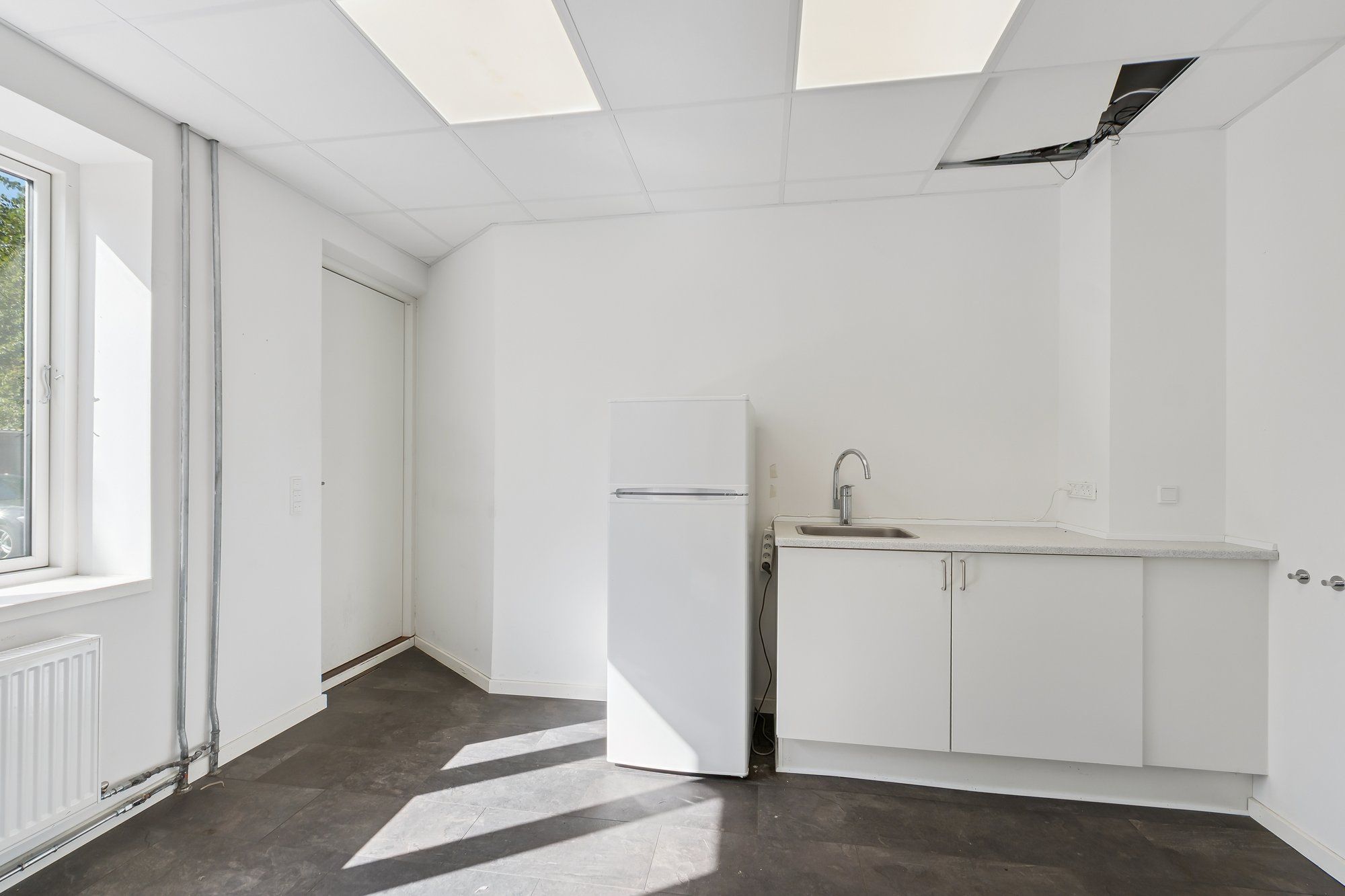 Rent og pænt kontor med lyse lofter og et lille køkkenhjørne med hvidt køleskab og en håndvask