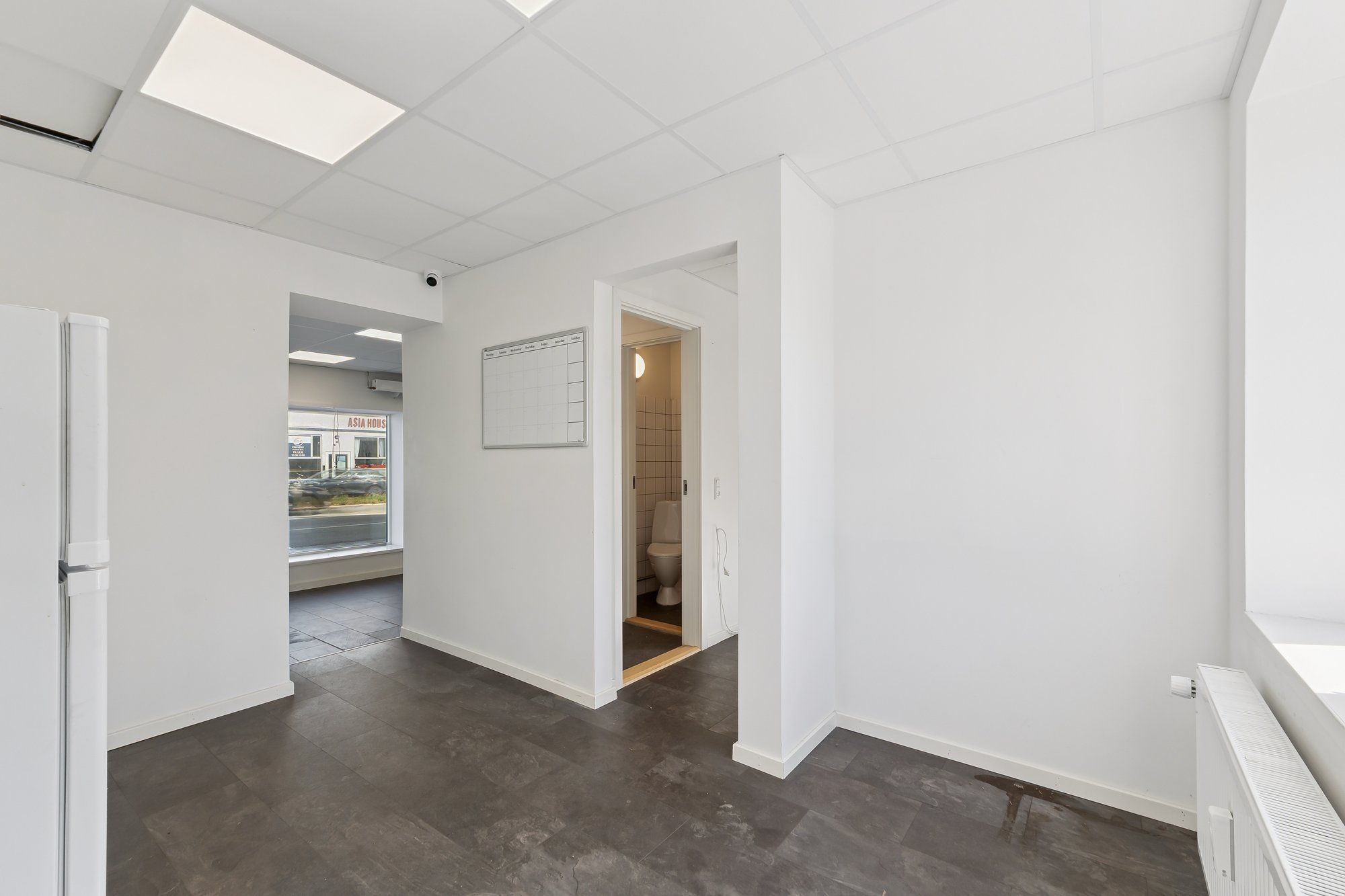 Store hvide vægge og lofter med to lyseplader. Et hvidt køleskab, en åben dør, et tomt badeværelse og en dør, der fører til