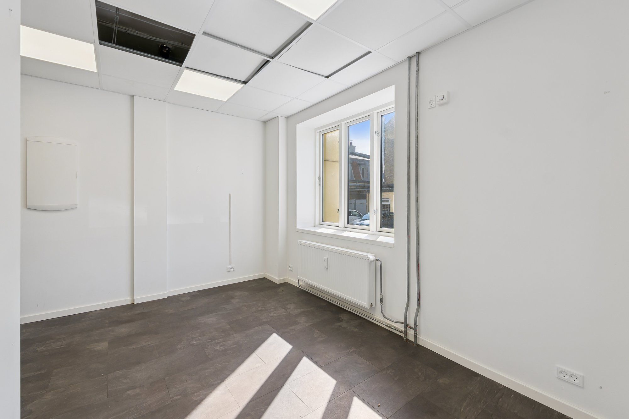 Et tomt rum med hvide vægge, gulvtæppe, radiator, vinduer og indbygget elektronisk udstyr