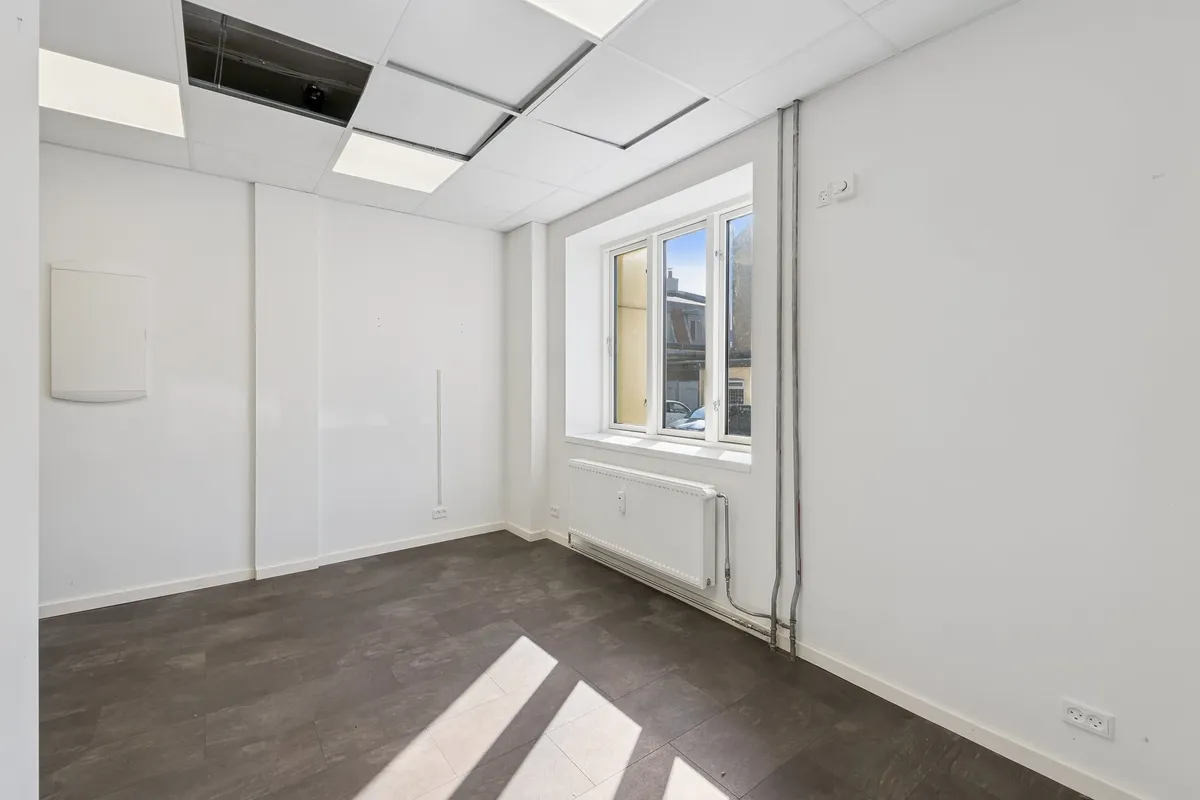 Et tomt rum med hvide vægge, gulvtæppe, radiator, vinduer og indbygget elektronisk udstyr