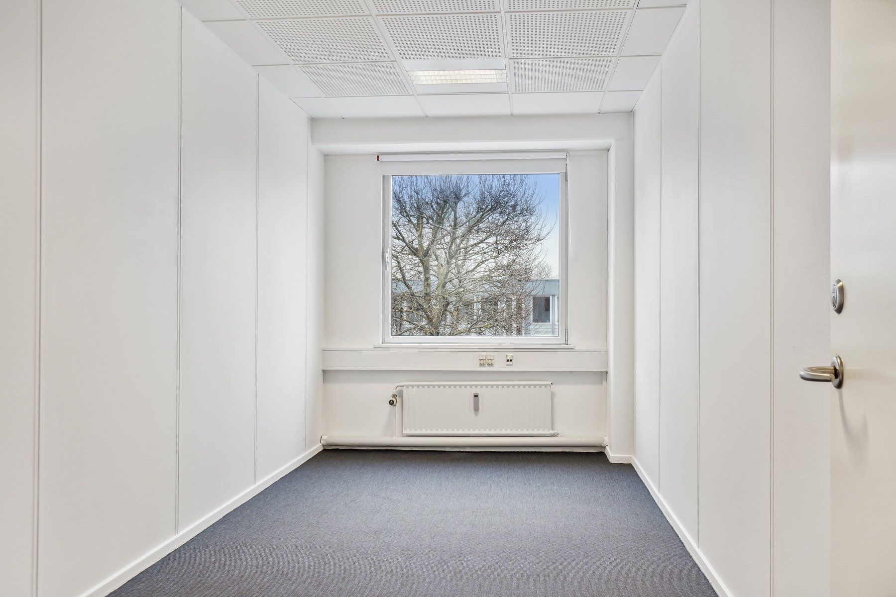 Et tomt kontorlokum med hvide vægge, højt til loftet og gulvbelægning i gråt. Der er en radiator under vinduet og et vindue