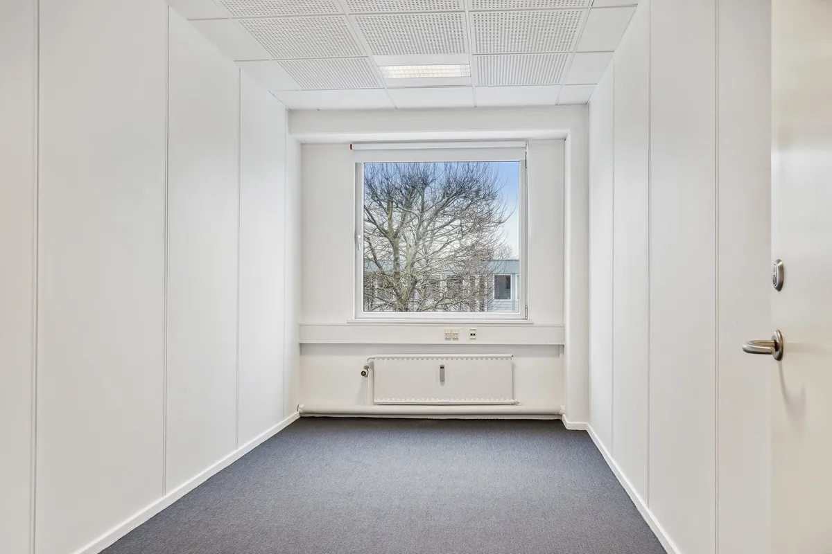 Et tomt kontorlokum med hvide vægge, højt til loftet og gulvbelægning i gråt. Der er en radiator under vinduet og et vindue