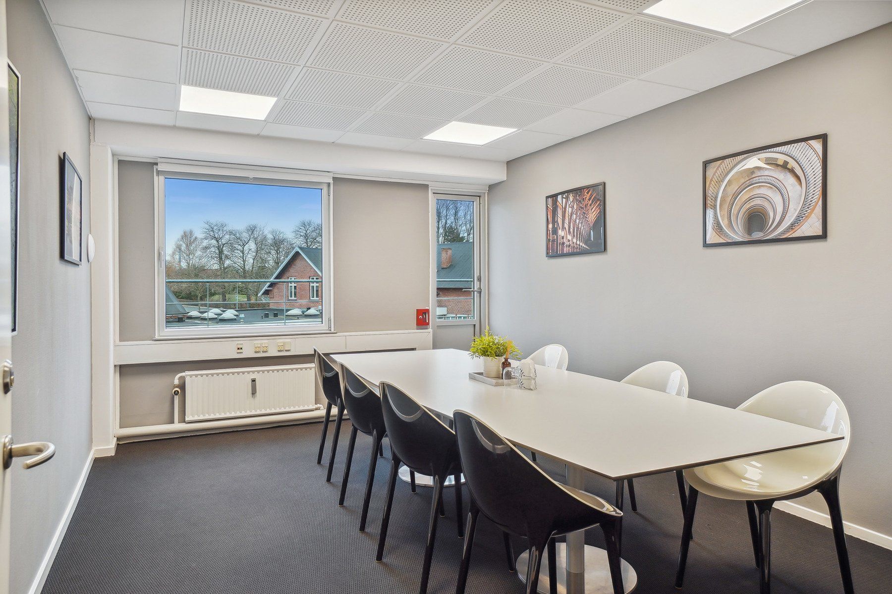 Kontor med langt, rektangulært bord, høje ruder, radiator og tre malerier med spiraler, spiraltrapper og kirke