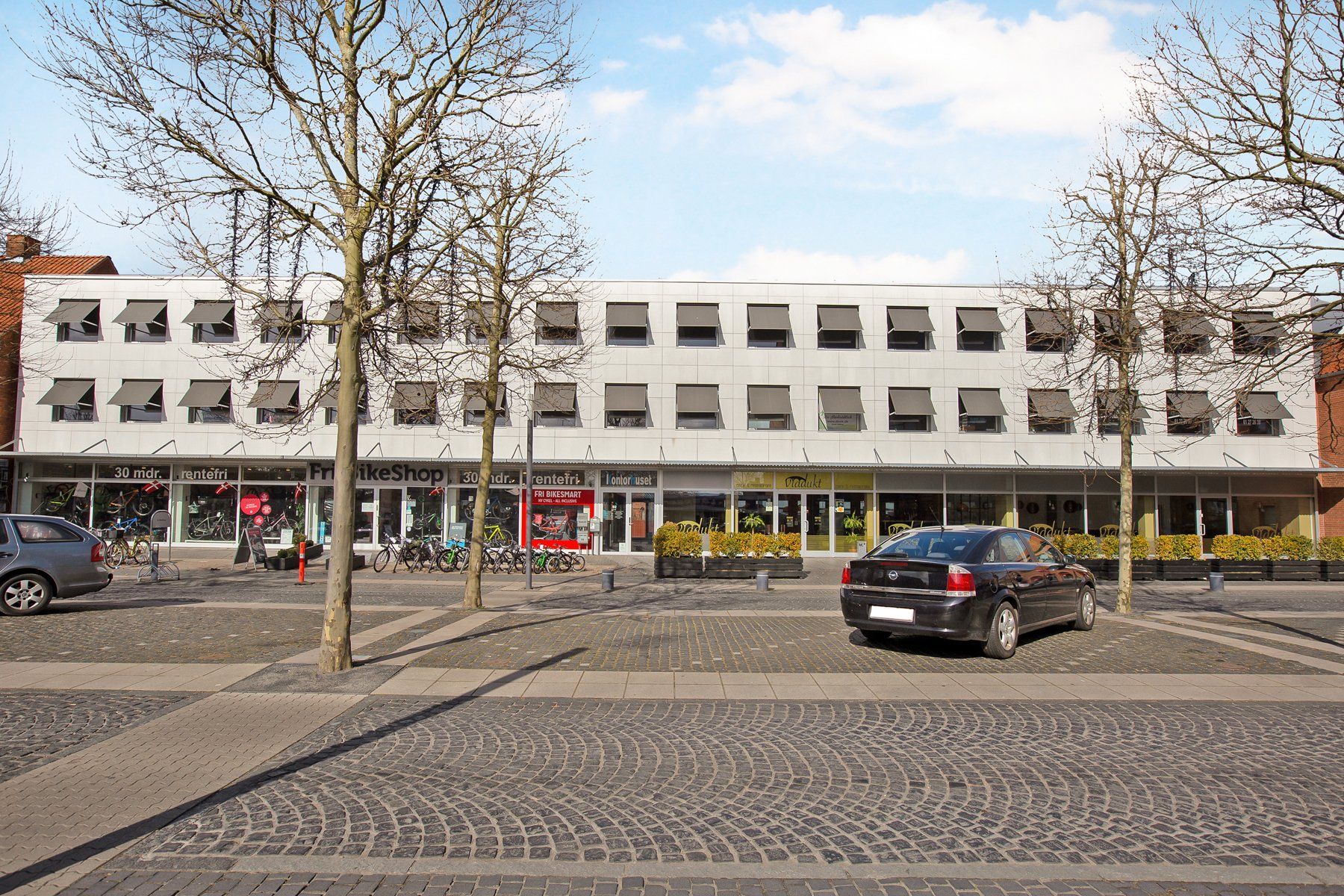 To biler parkeret ved et butikscenter med forretninger og træer og enten fritid eller cykeludlejning