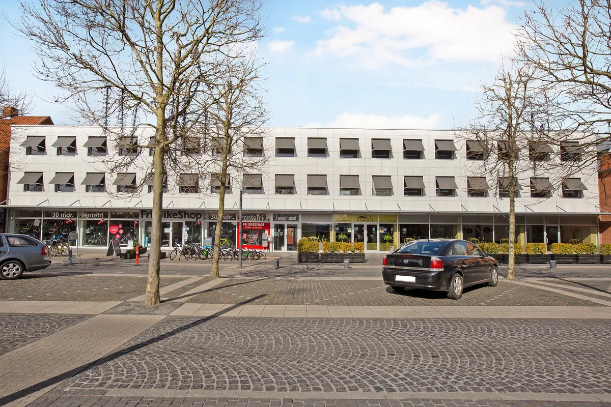 To biler parkeret ved et butikscenter med forretninger og træer og enten fritid eller cykeludlejning