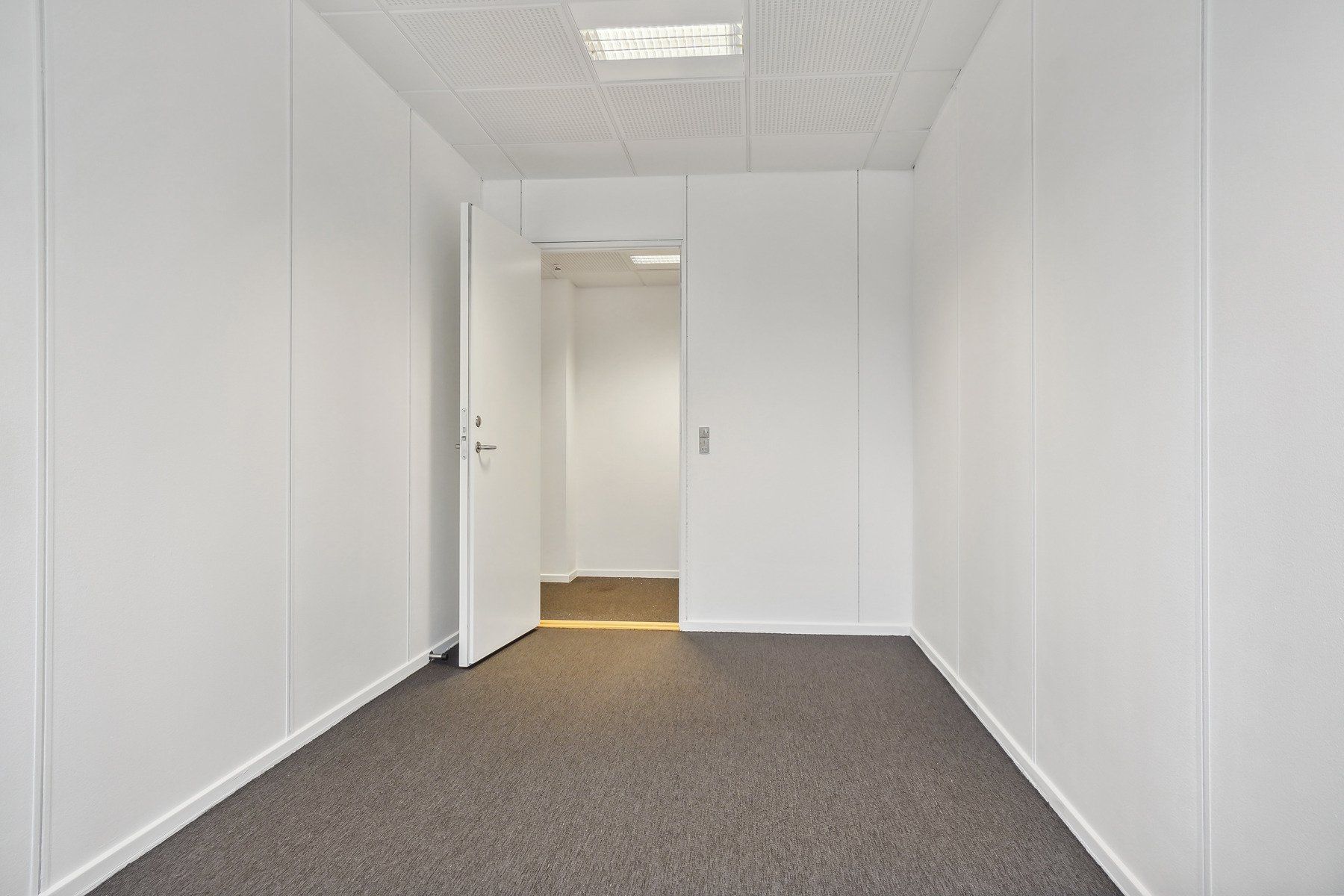 En lang og smal korridor med hvide vægge og en åben dør, der fører til en anden rummelighed med lysegul gulvbelægning og
