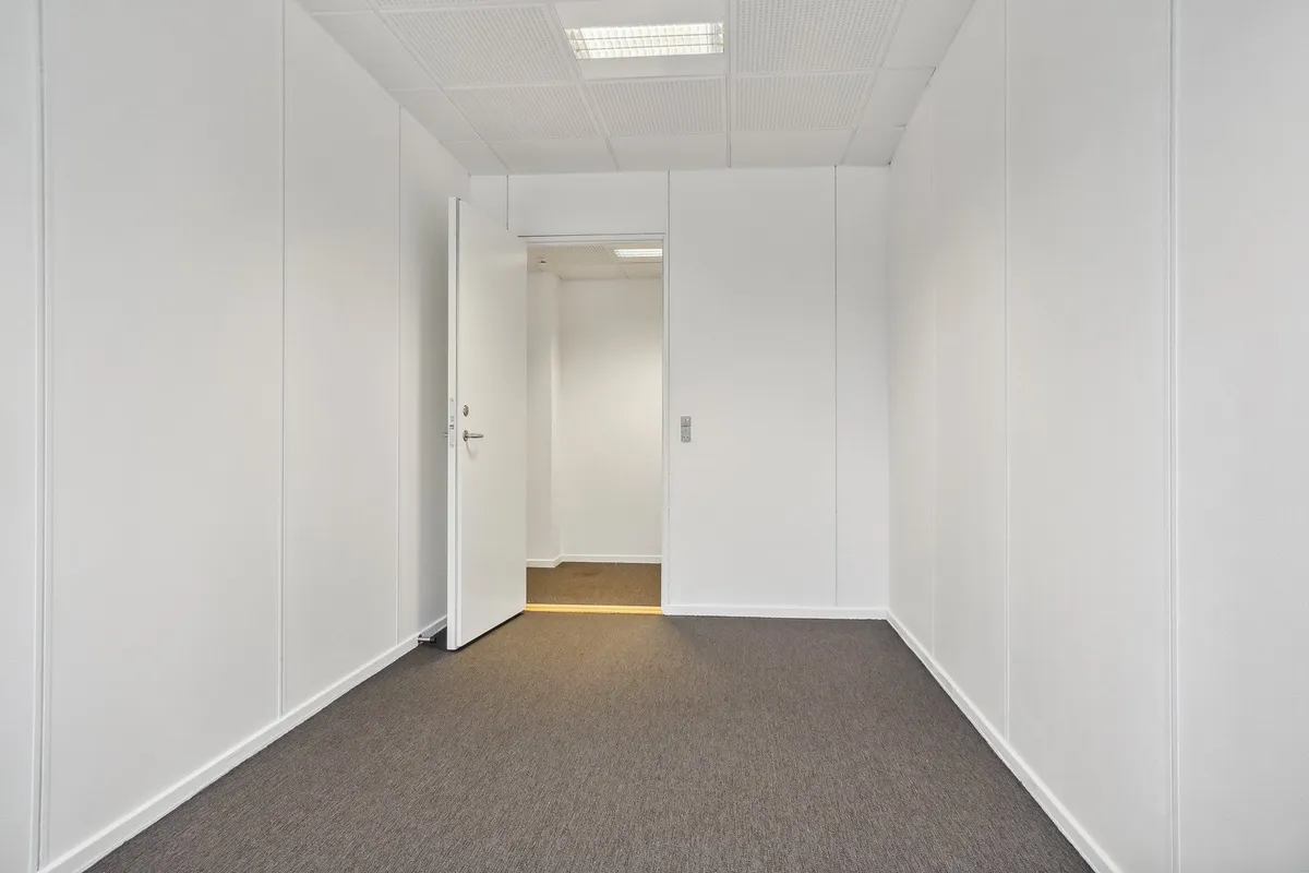 En lang og smal korridor med hvide vægge og en åben dør, der fører til en anden rummelighed med lysegul gulvbelægning og