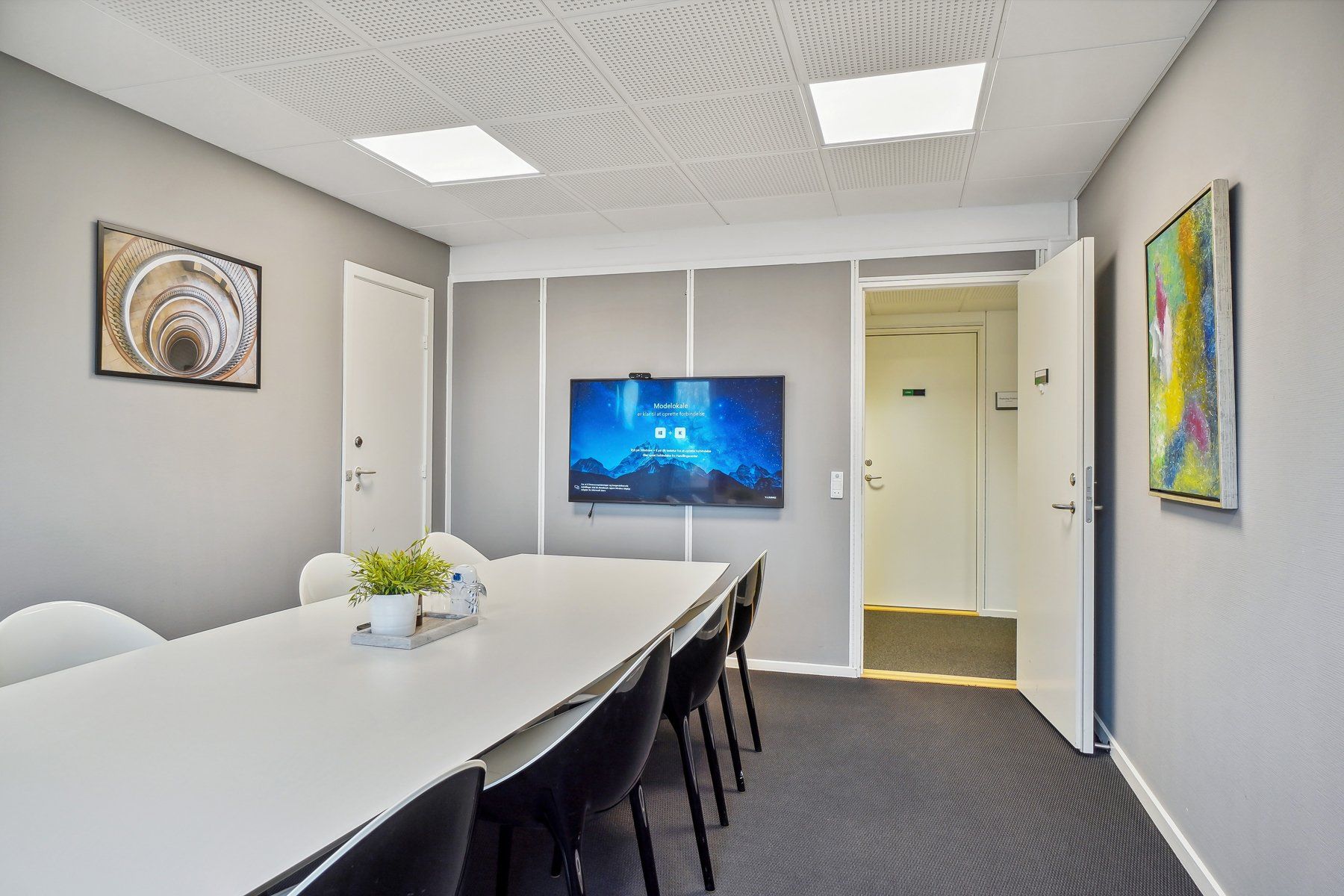 Kontor med bord, stole, plante, tv-skærm, åben dør og malerier på væggen