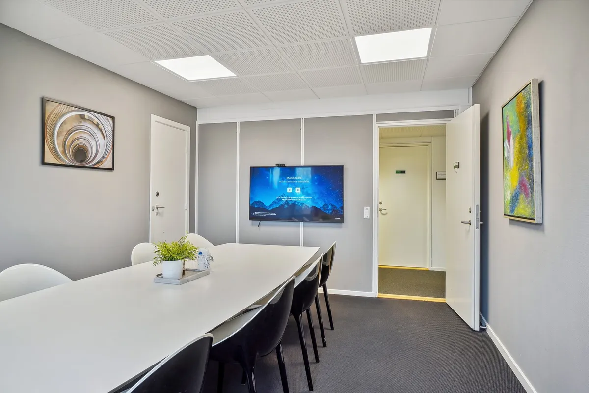 Kontor med bord, stole, plante, tv-skærm, åben dør og malerier på væggen