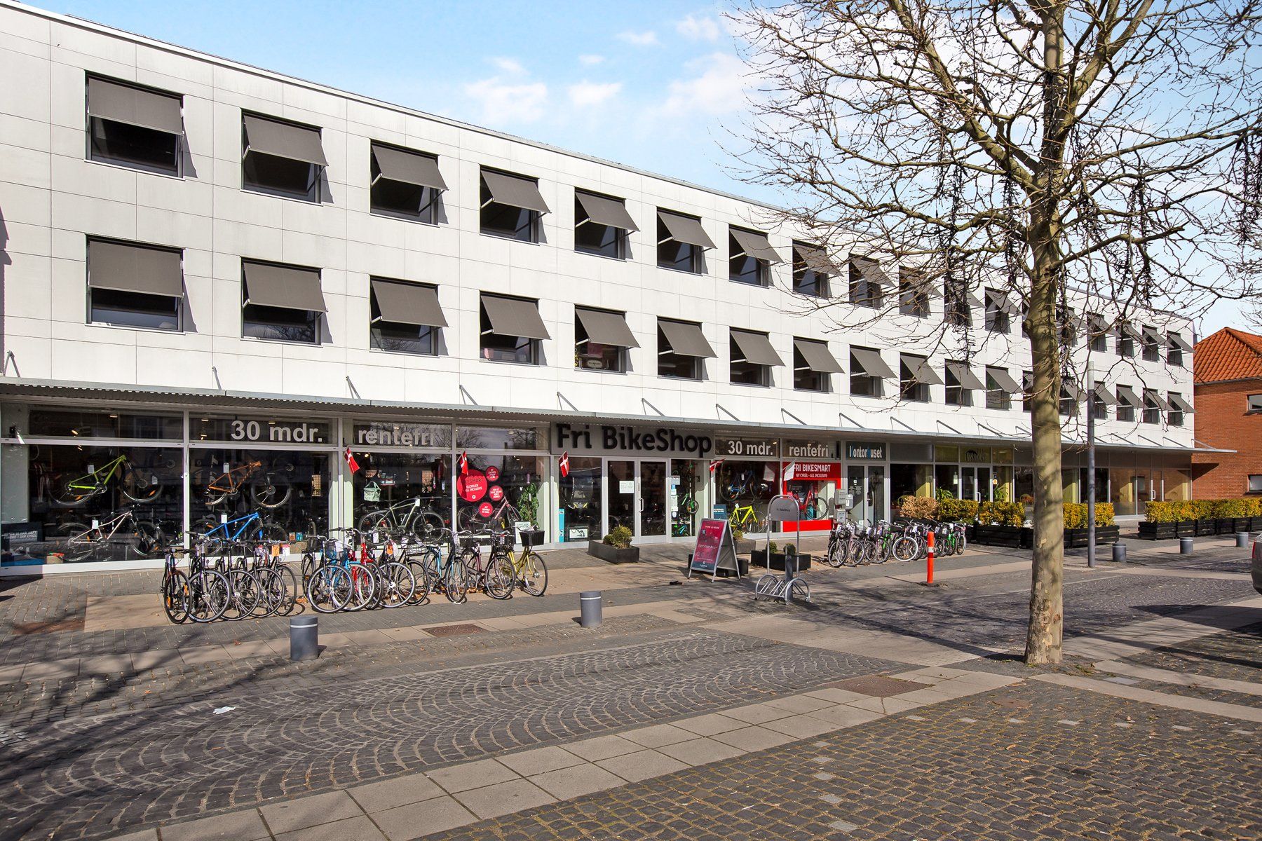 Fri BikeShop med mange lejecykler parkeret foran og vinduer med solskærme og flag i Taastrup