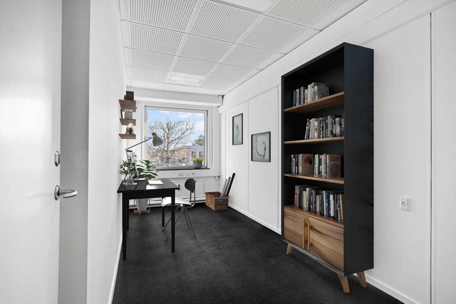 Kontor med sort bord og lampe, to stole, høj boghylde med bøger, vindue og plante. Et hjørne af et kontorfællesskab med