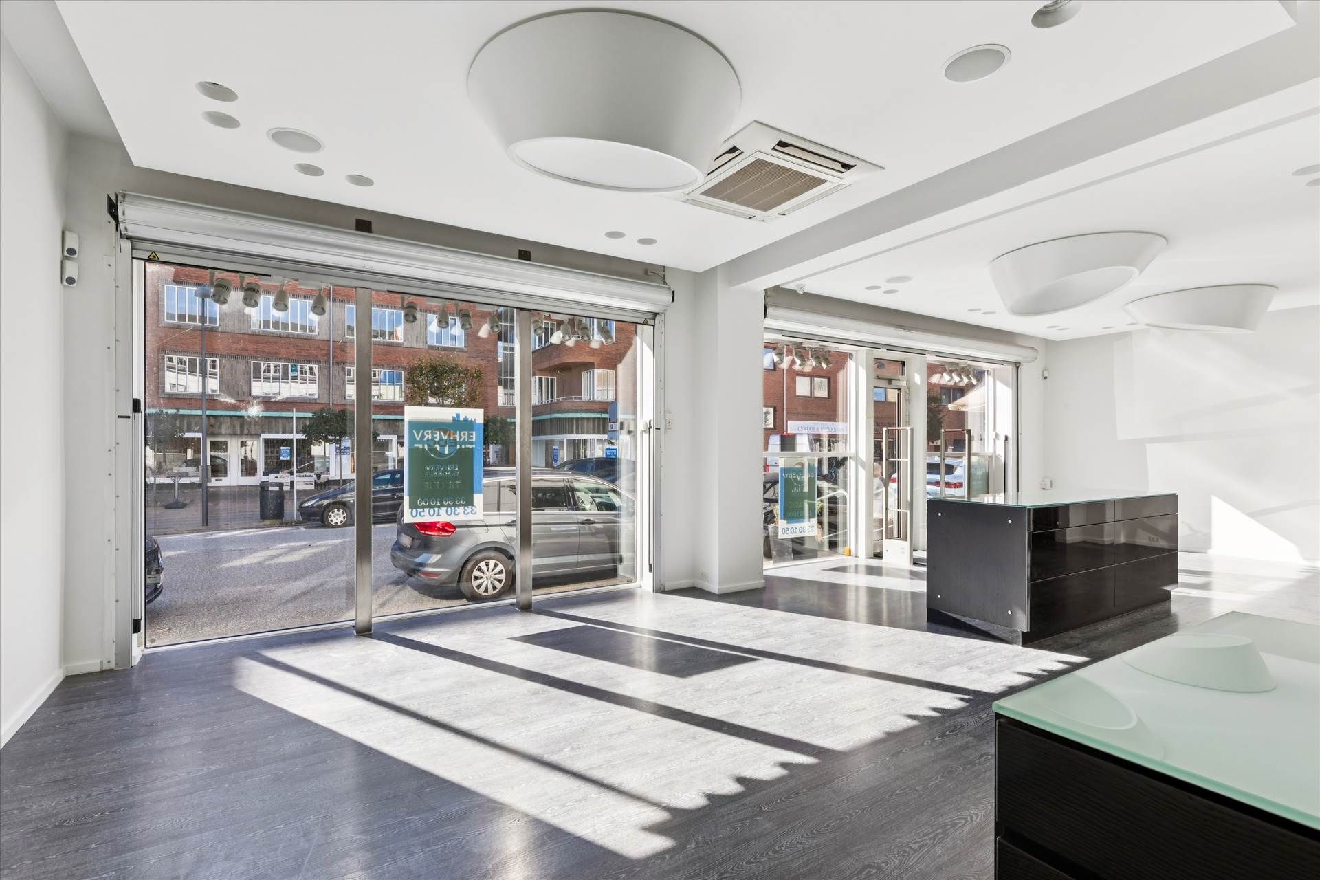 En butikslokal med store glasdøre og lysegulve samt en mørk skranken og en gulvmonteret airconditioner