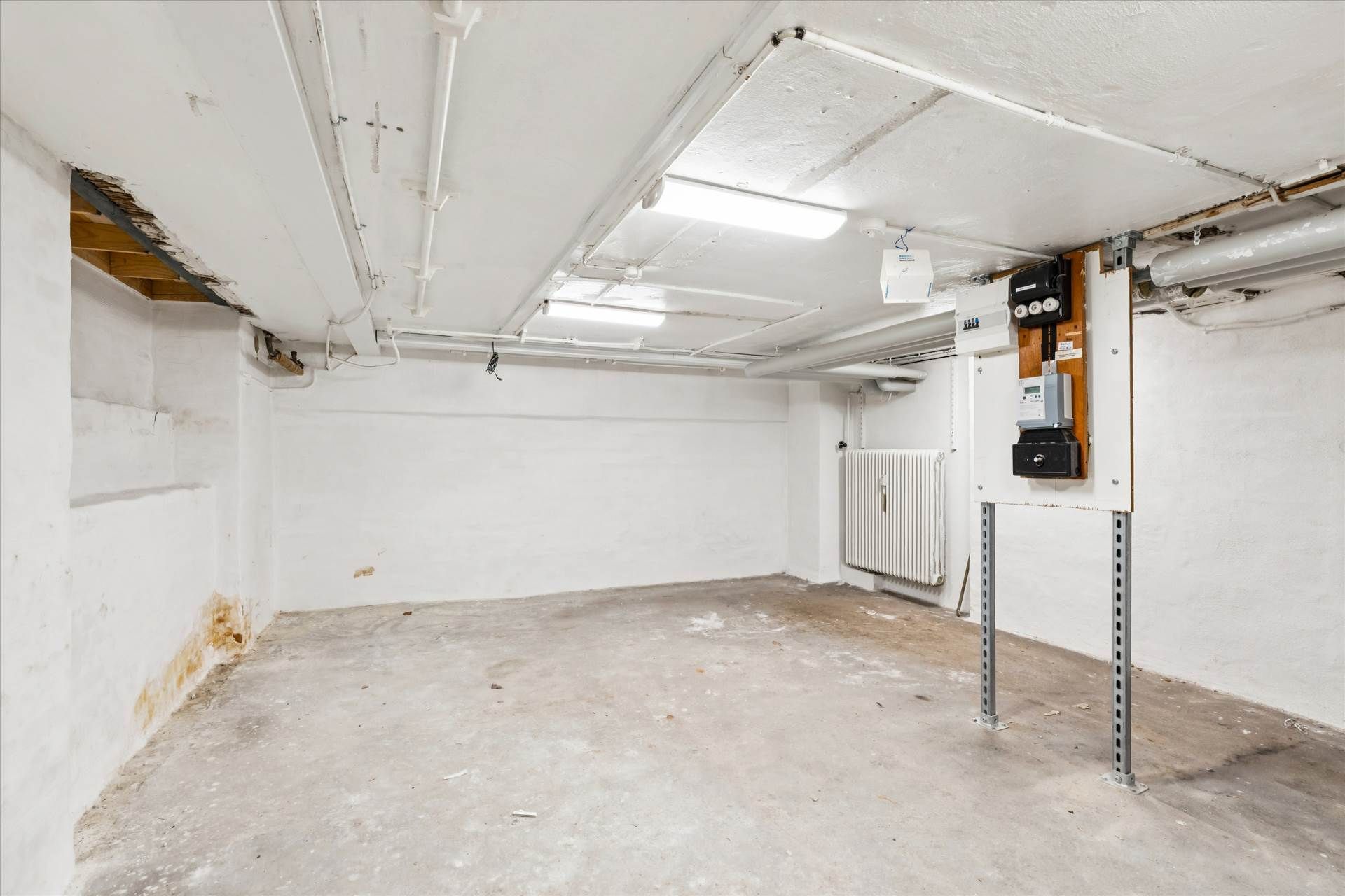 En stor, ubenyttet lagerhal med hvide vægge og cementgulv, hvor der er lysekroner og rør i loftet og et hvidt radiator på