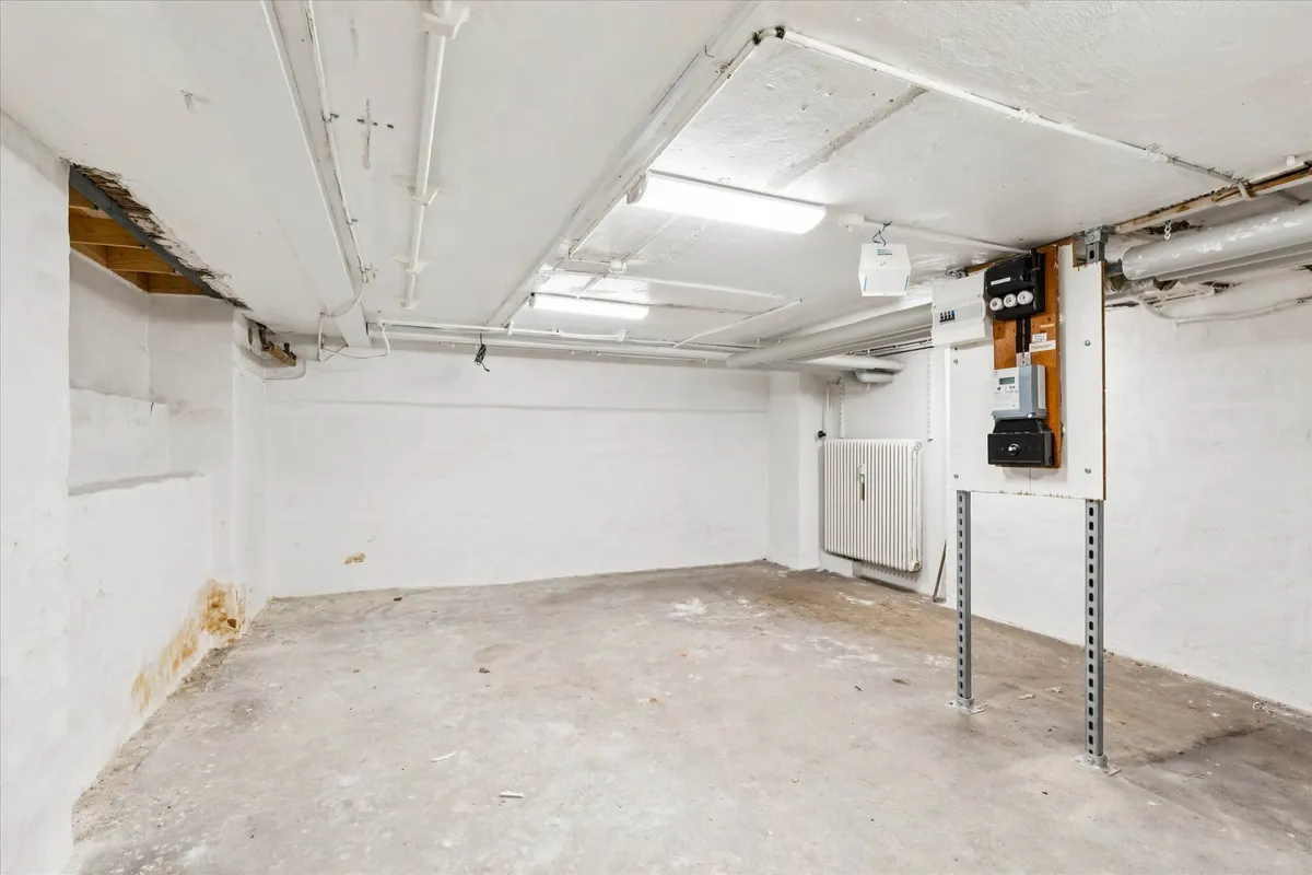 En stor, ubenyttet lagerhal med hvide vægge og cementgulv, hvor der er lysekroner og rør i loftet og et hvidt radiator på