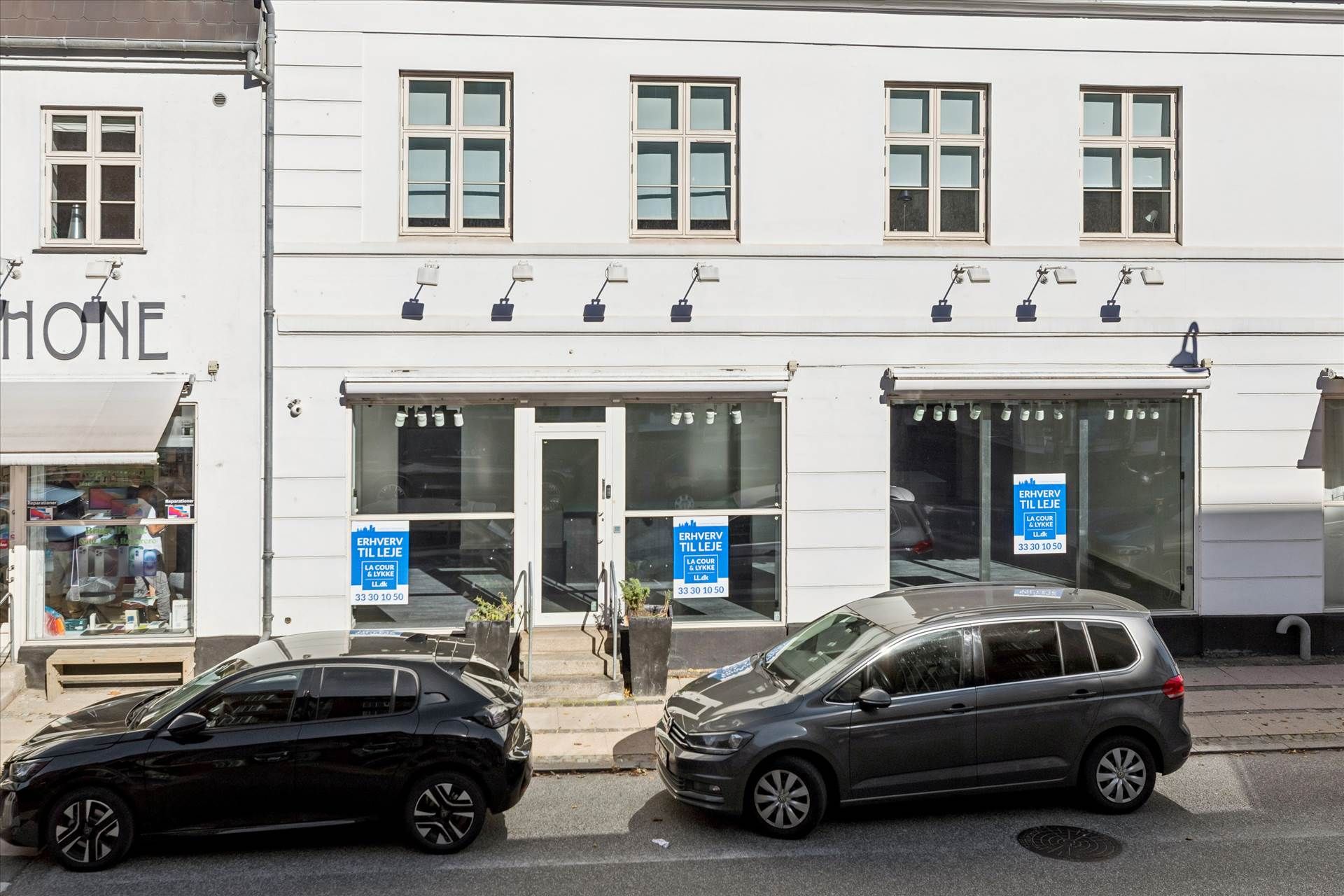 To biler parkeret foran en butik med reklameplakat og vinduer, der viser møbler og andre varer i butikken