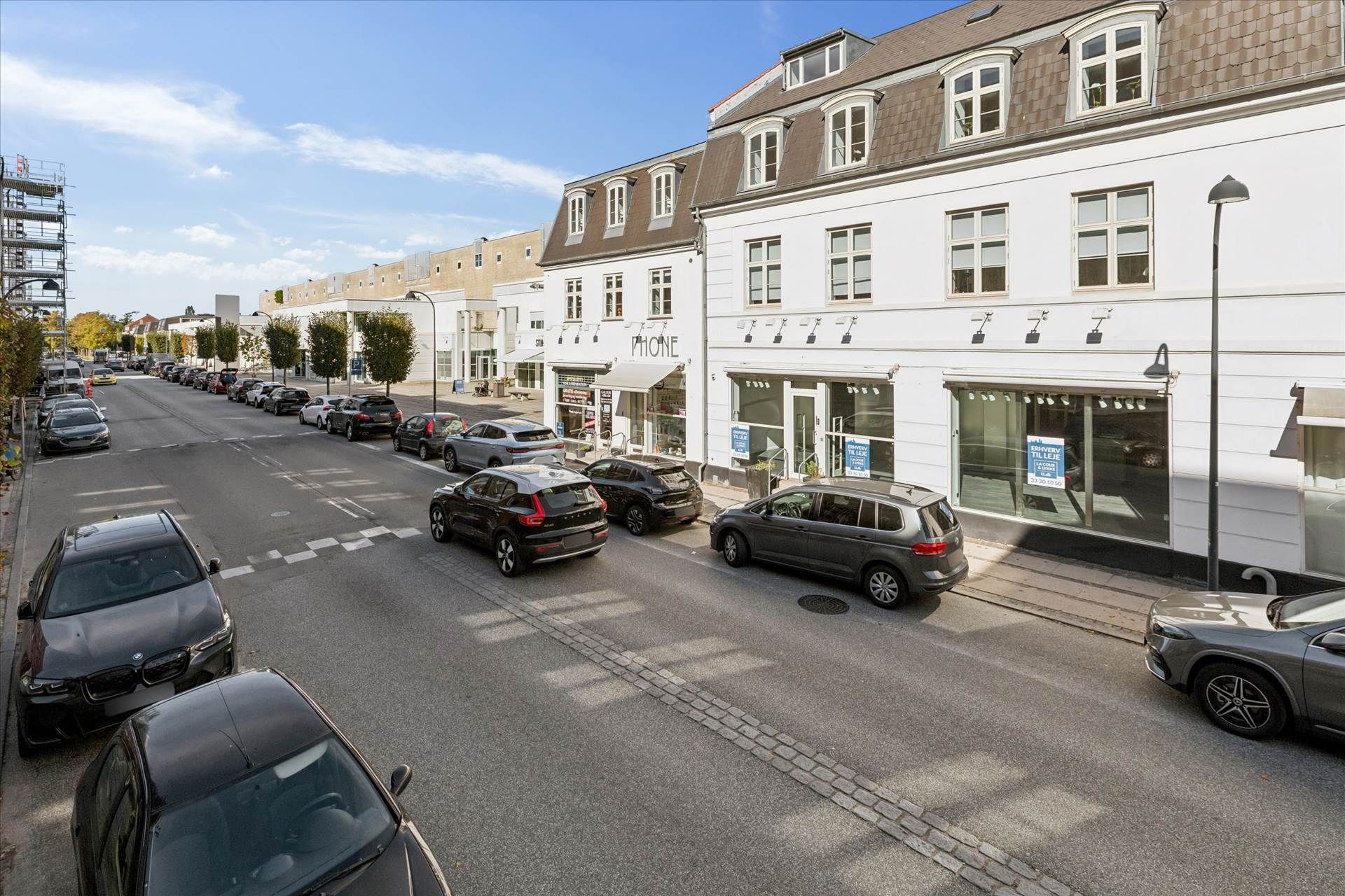 En bilparkeret gade med forretninger og biler på hver side, udsmykket med grønne planter og lysekroner