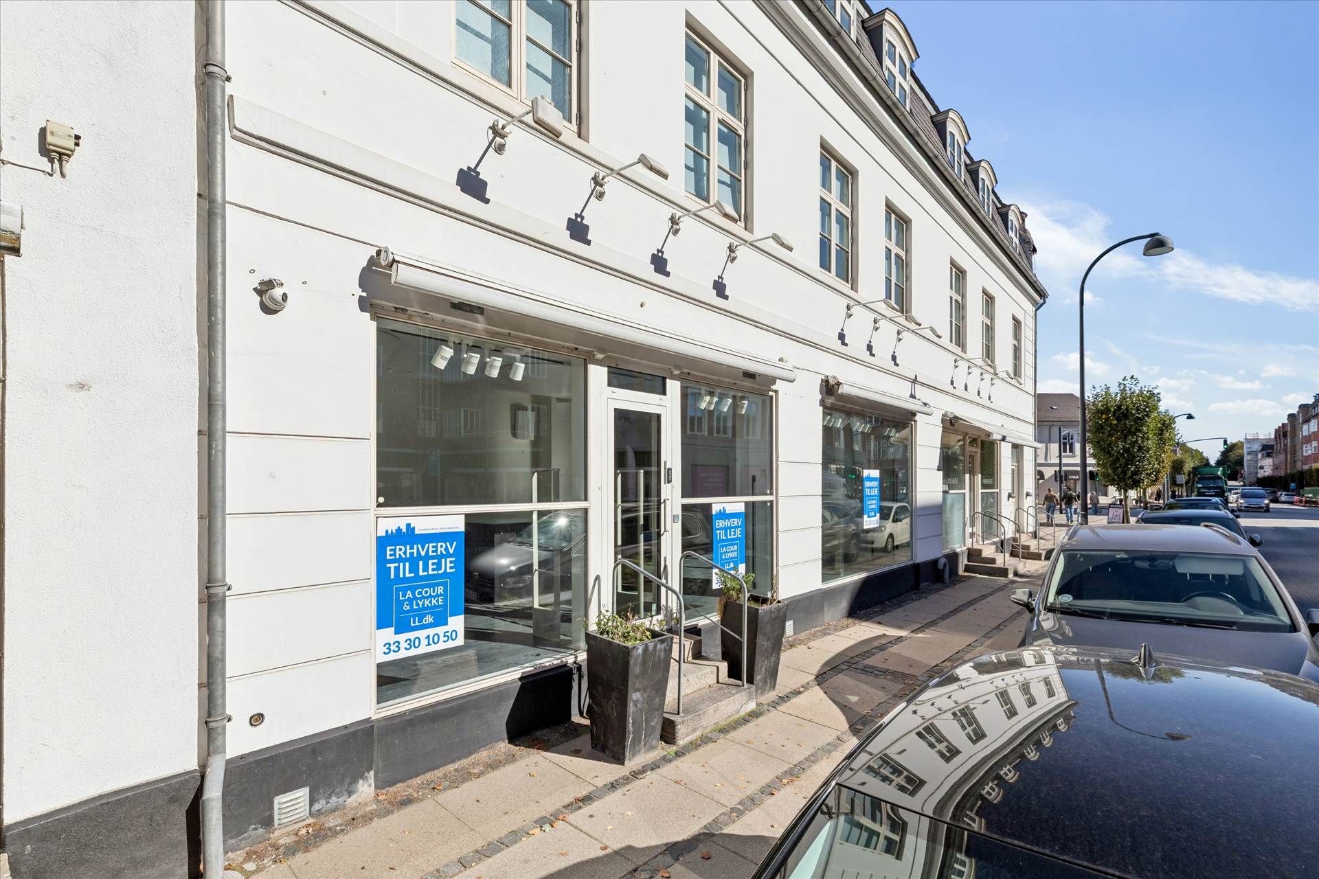 Bygning med vinduer og facader er reklamerede med tekst på dansk, og der er to planter og flere biler parkeret langs