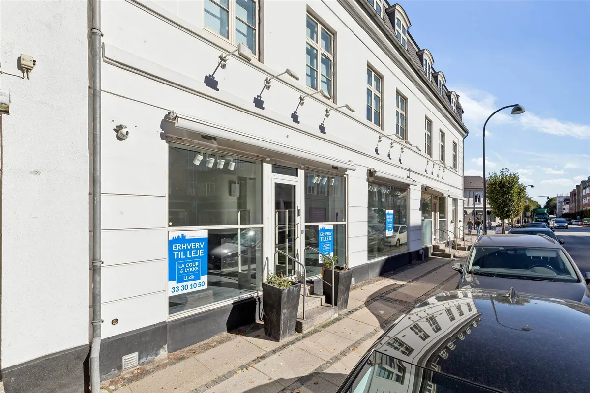 Bygning med vinduer og facader er reklamerede med tekst på dansk, og der er to planter og flere biler parkeret langs