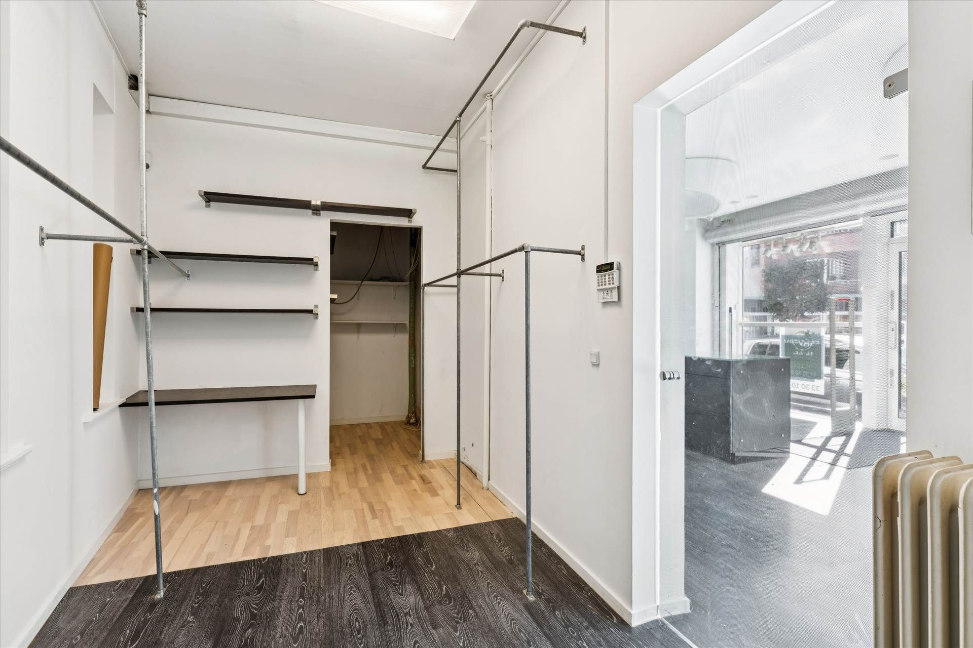 Indenfor et lokale med hvide vægge, trægulv og en stor glasdør er der et rum med metalhylder, hængeklæder og et gulvtæppe