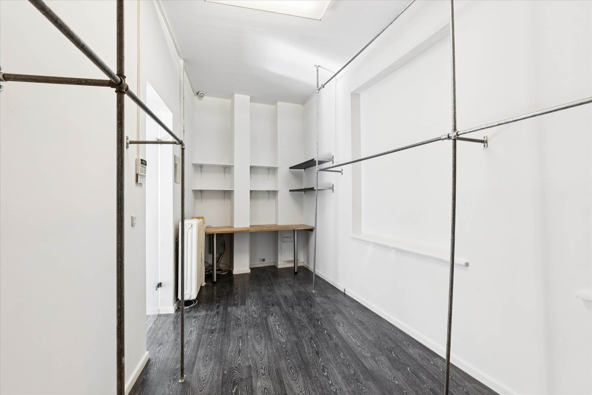 Indeni et minimalistisk og rensket værelse med hvide vægge og en mørk trægulv samt metalliske garderobebøjler